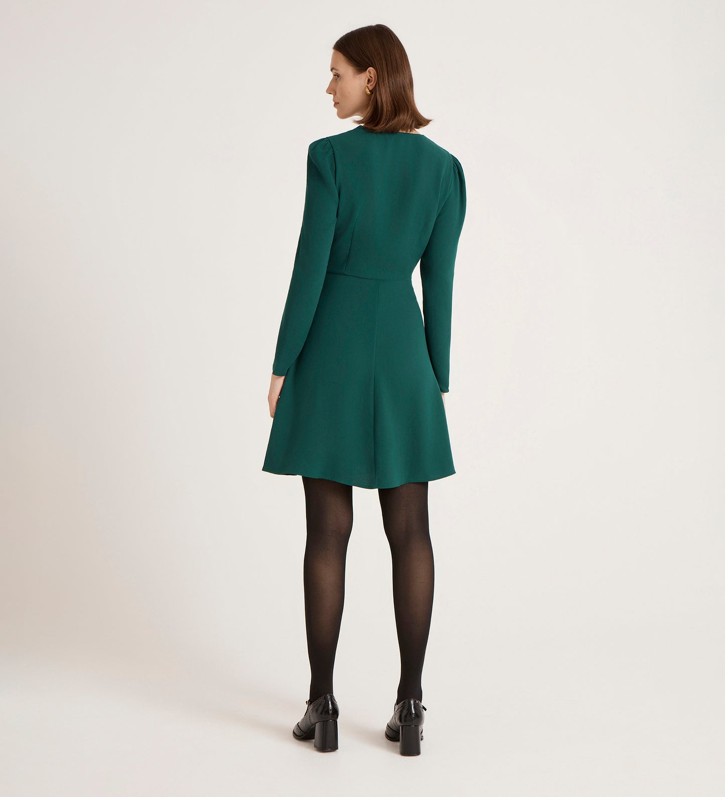 A woman with chin-length brown hair faces away, wearing the Kadie Bottle Green Crepe Short Dress with black tights and black heeled shoes against a plain light background.
