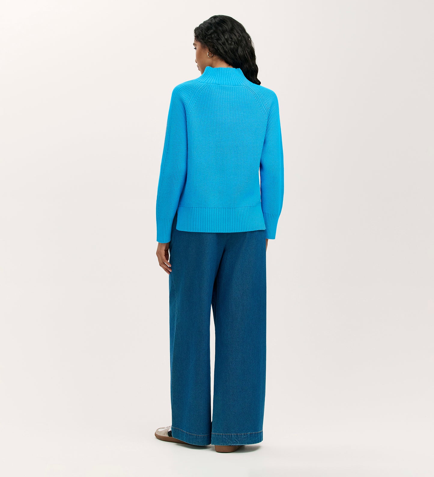 A person with long, wavy hair faces away, wearing the Kaela Light Blue High Neck Jumper and wide-leg blue jeans, paired with brown shoes, against a plain light background for an effortlessly stylish look.