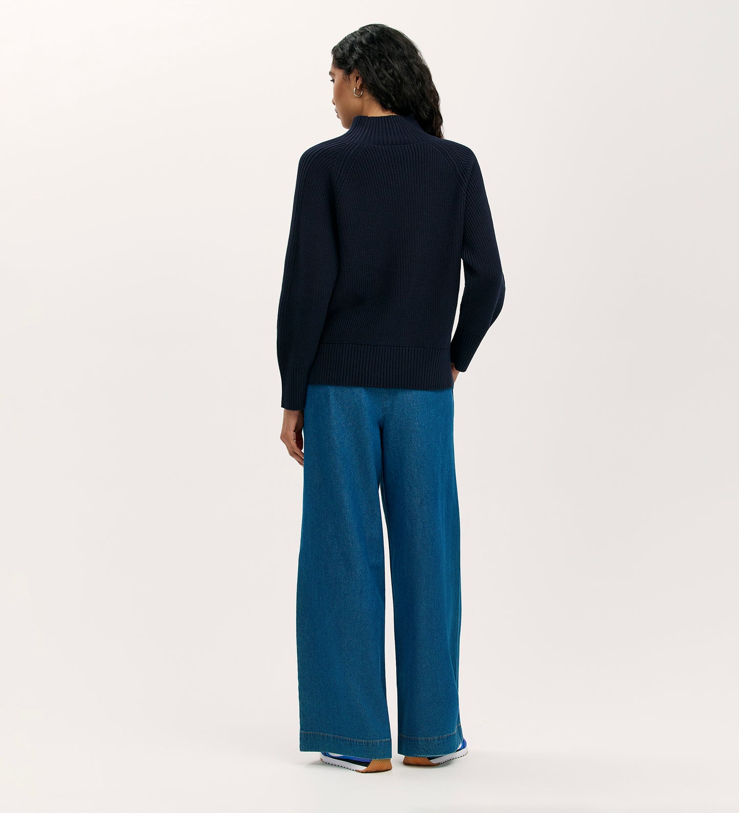 A person with long dark hair stands facing away in the Kaela Navy High Neck Jumper, wide-leg blue jeans, and white sneakers against a plain light background.