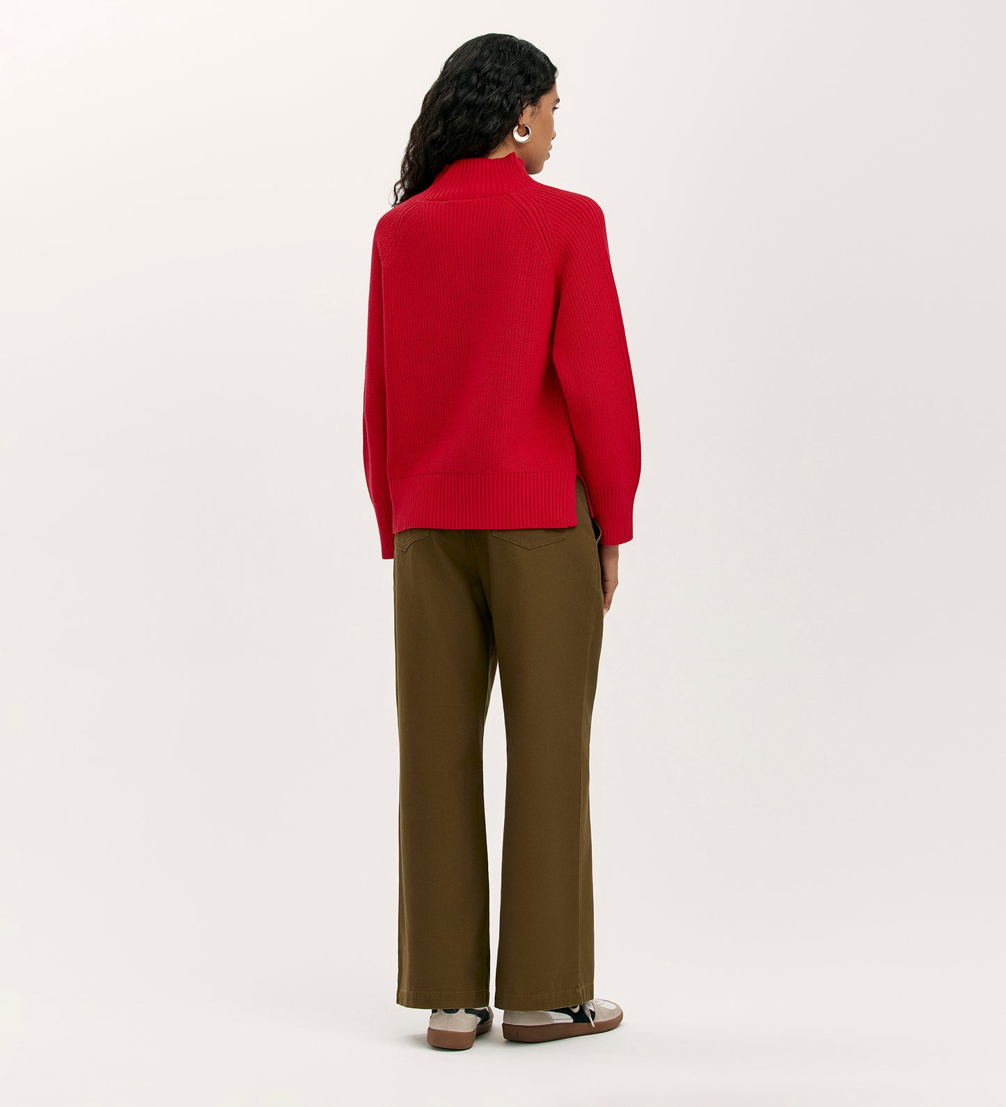 A person with long, dark, wavy hair stands facing away in the Kaela Red High Neck Jumper and olive green wide-leg pants, paired with beige shoes—showcasing a women’s wardrobe staple against a plain light background.