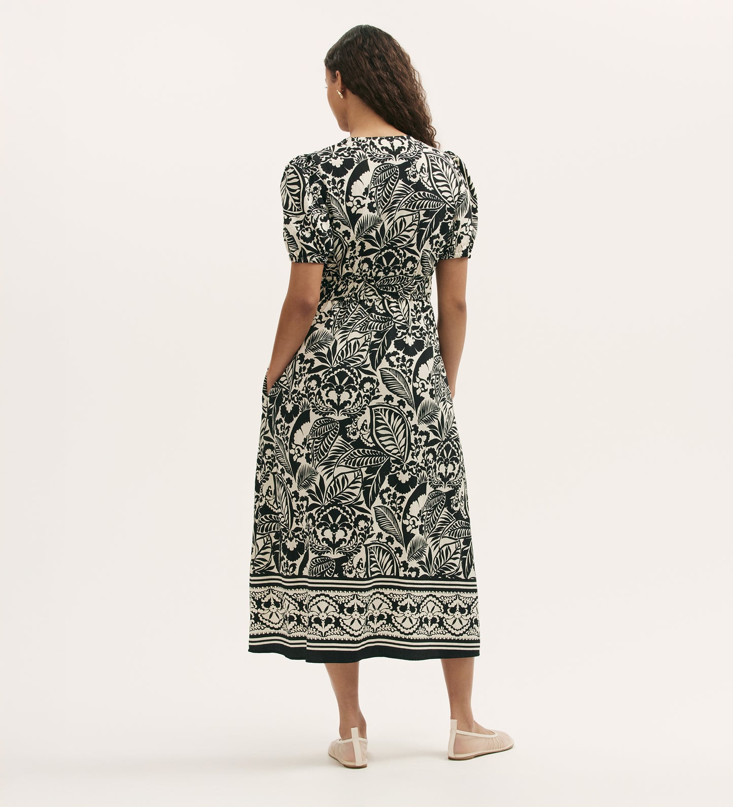 A woman with loose curly hair stands facing away, wearing the Katerina Black Border Leaf Crepe Midi Smock Dress featuring a black and white leaf print, border hem design, and cream flat shoes against a plain background.