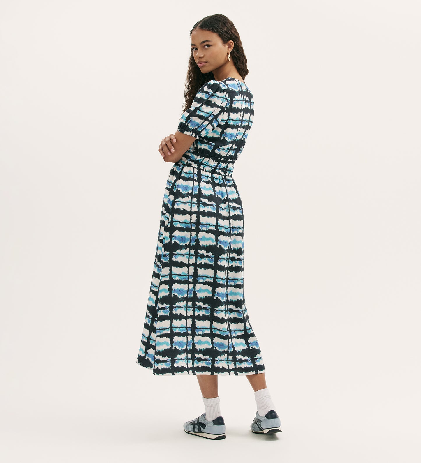 A woman stands sideways, looking over her shoulder, wearing the Katerina Blue Tie Dye Midi Smock Dress with short sleeves and an elasticated waistband, paired with sneakers and white socks. The background is plain white.