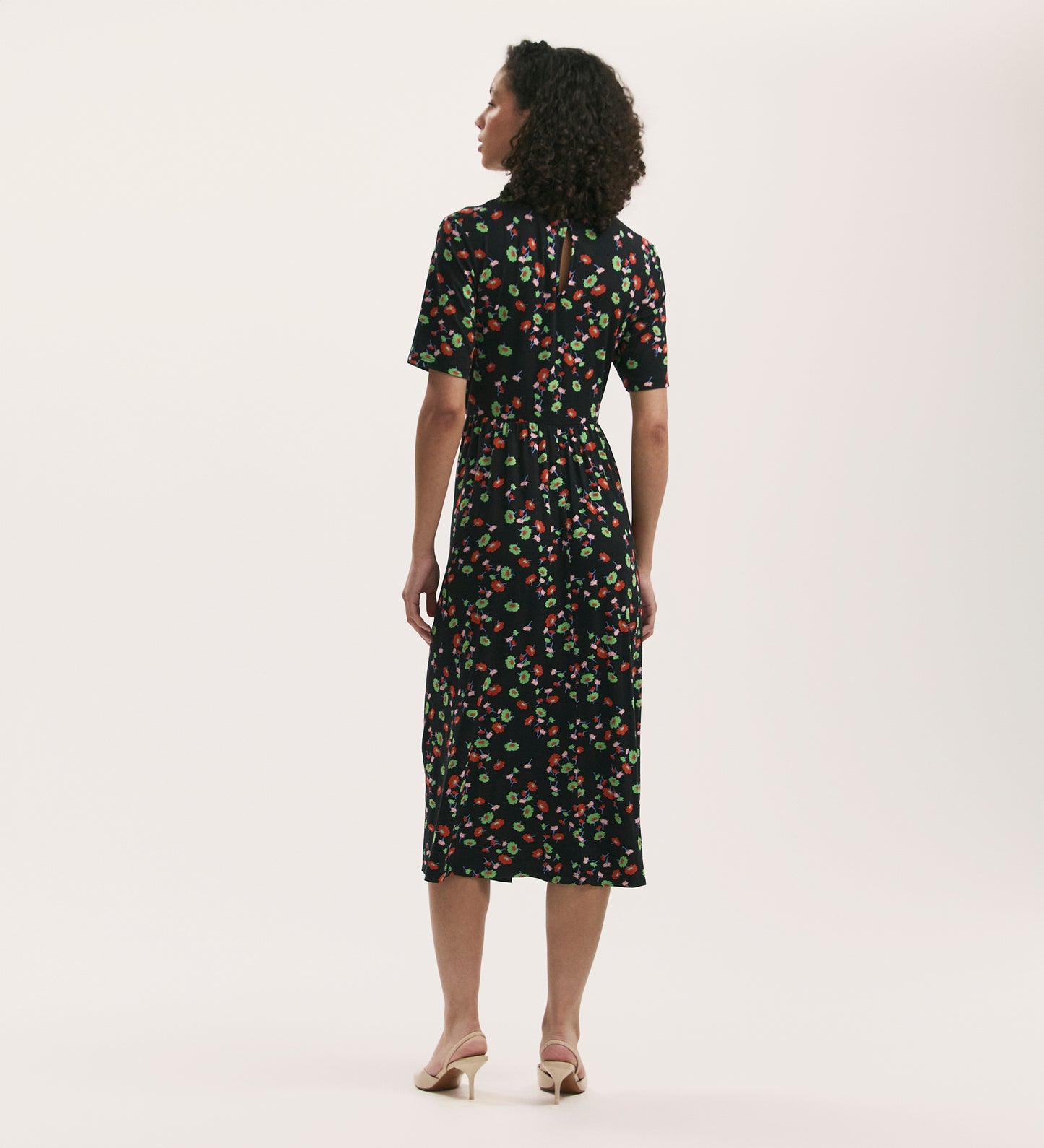 A woman with curly hair faces away from the camera, wearing the Kathryn Black Flower Jersey Midi T-Shirt Dress. The soft jersey fabric and relaxed fit flatter her silhouette. She pairs it with light heeled sandals against a plain background.