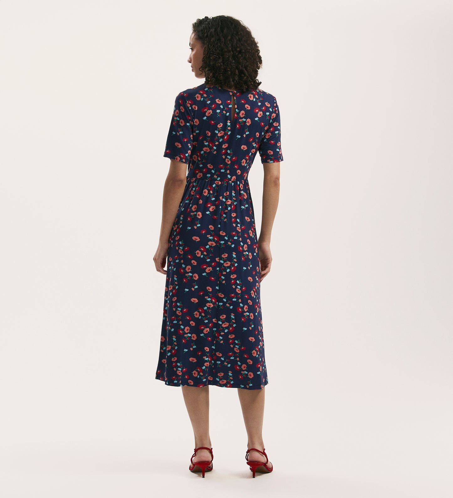 A woman with curly hair stands facing away, wearing the Kathryn Navy Flower Jersey Midi T-Shirt Dress and red heeled sandals against a plain white background.