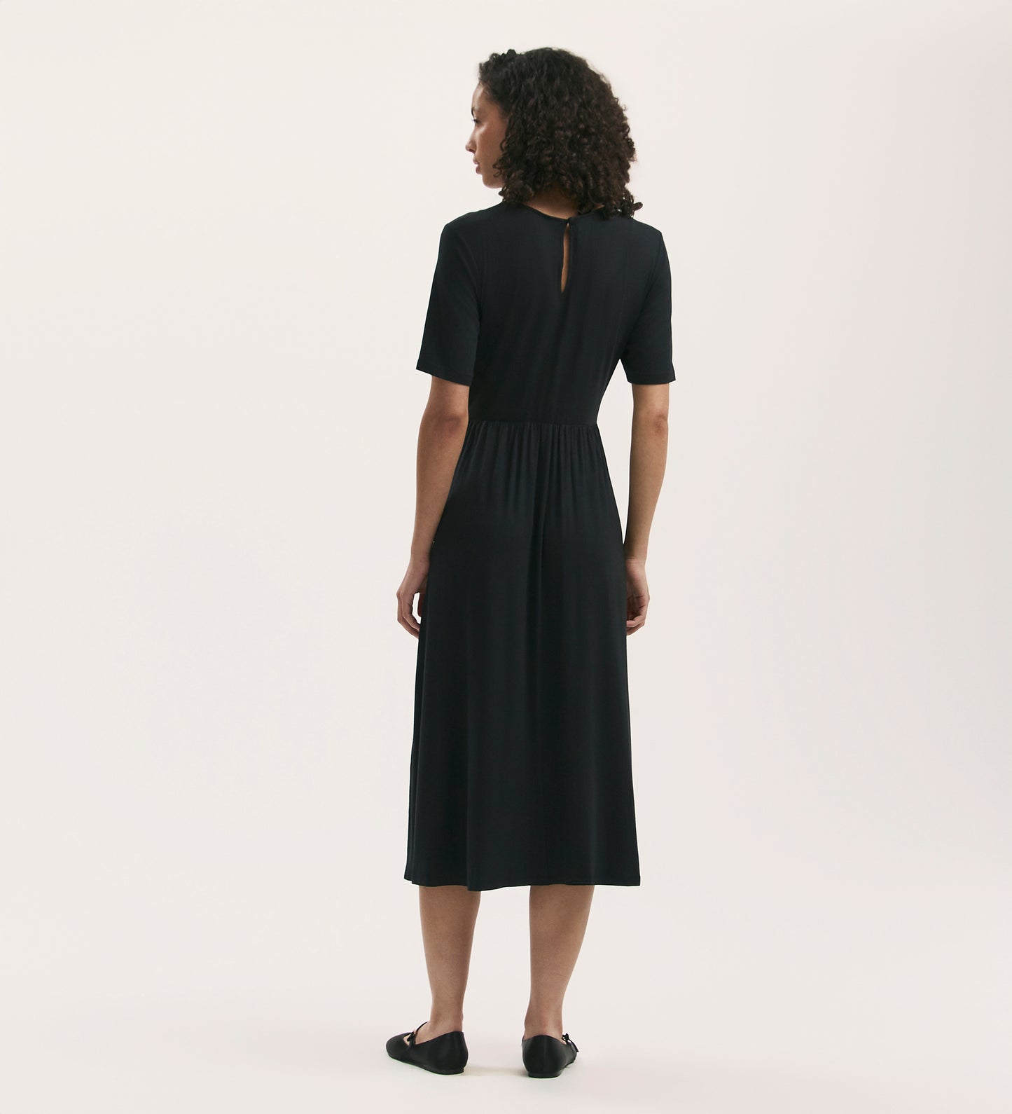 A woman with curly hair faces away, wearing the Kathryn Black Jersey Midi T-Shirt Dress—relaxed fit, soft jersey, short sleeves, keyhole back—paired with black flats against a plain light background.