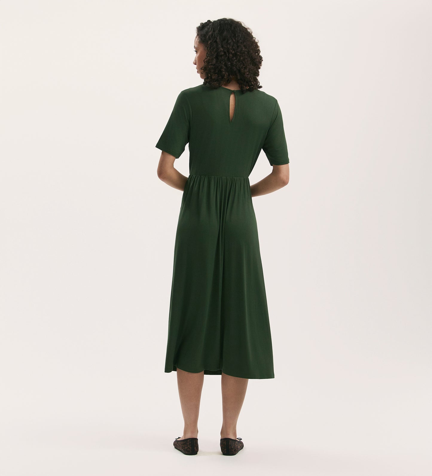 A woman with curly hair faces away, wearing the Kathryn Khaki Jersey Midi T-Shirt Dress featuring a gathered waist and keyhole back detail. She pairs it with black flats, set against a plain light background.
