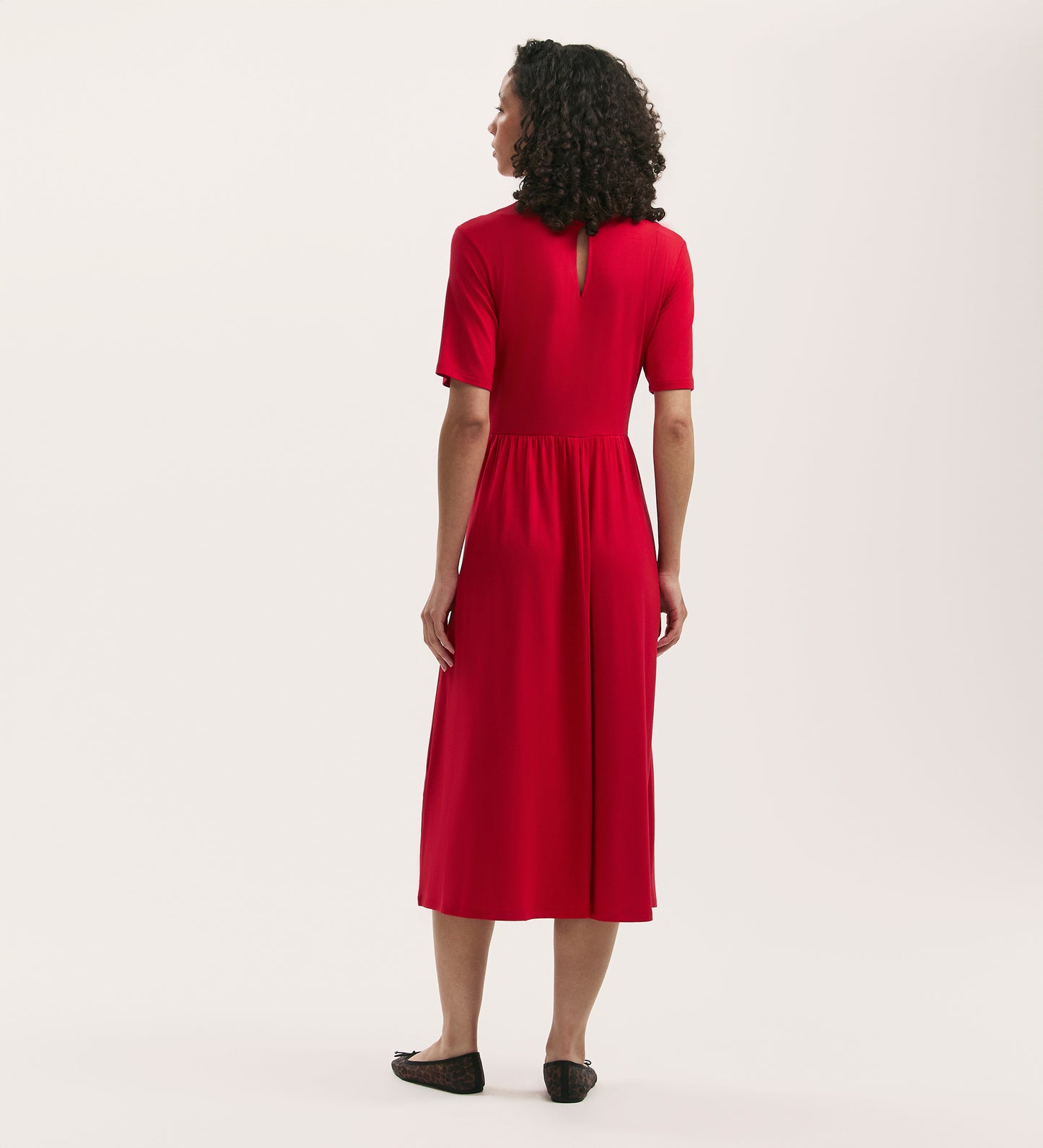 A woman with curly hair faces away, wearing the Kathryn Red Jersey Midi T-Shirt Dress—relaxed fit, soft jersey fabric—with black flat shoes against a plain light background.