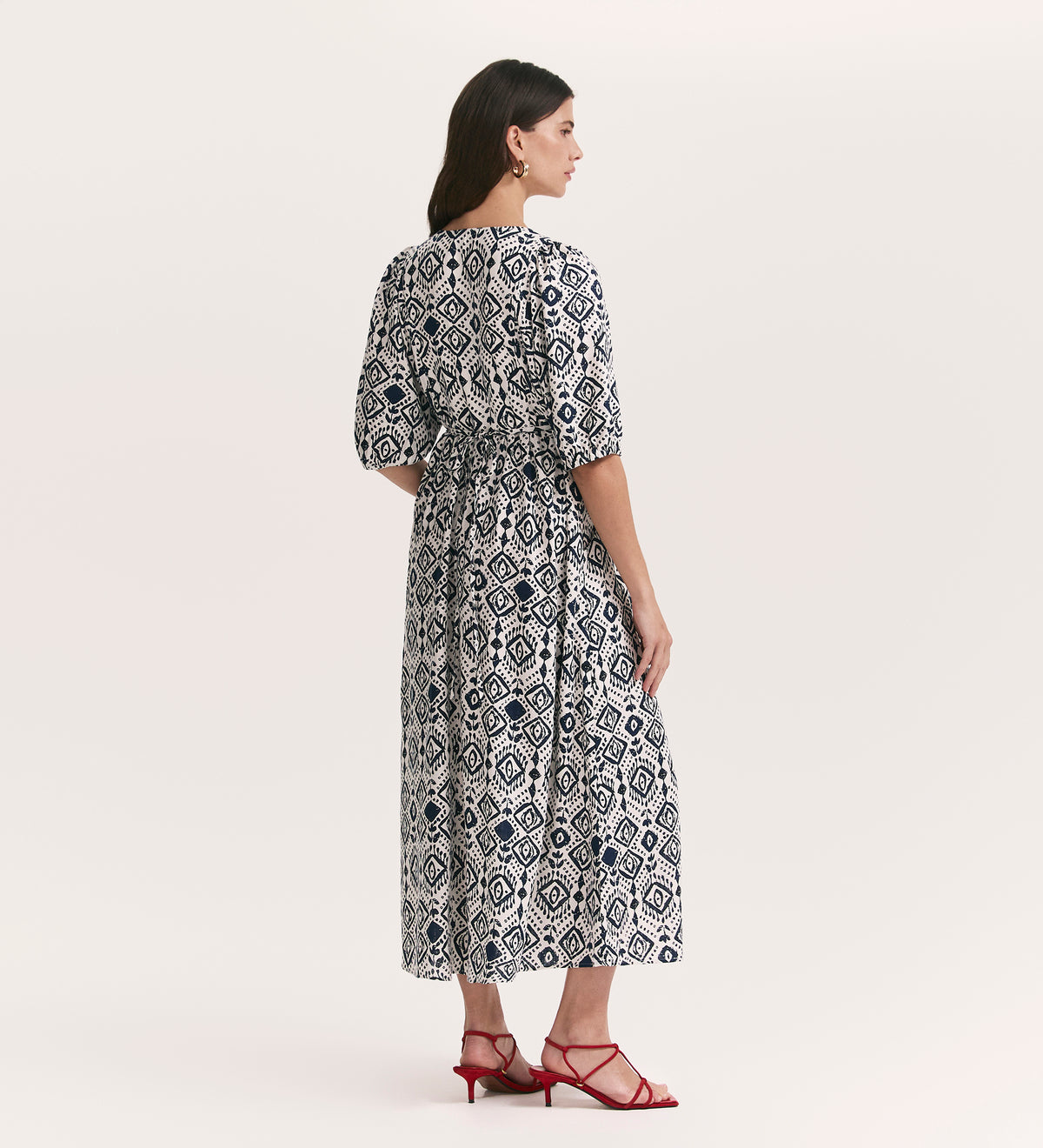 A woman stands facing away in the Katie Ivory Woodblock Midi Smock Dress with Linen, featuring elbow-length sleeves and a black geometric print. She pairs it with red strappy sandals against a plain, light background.