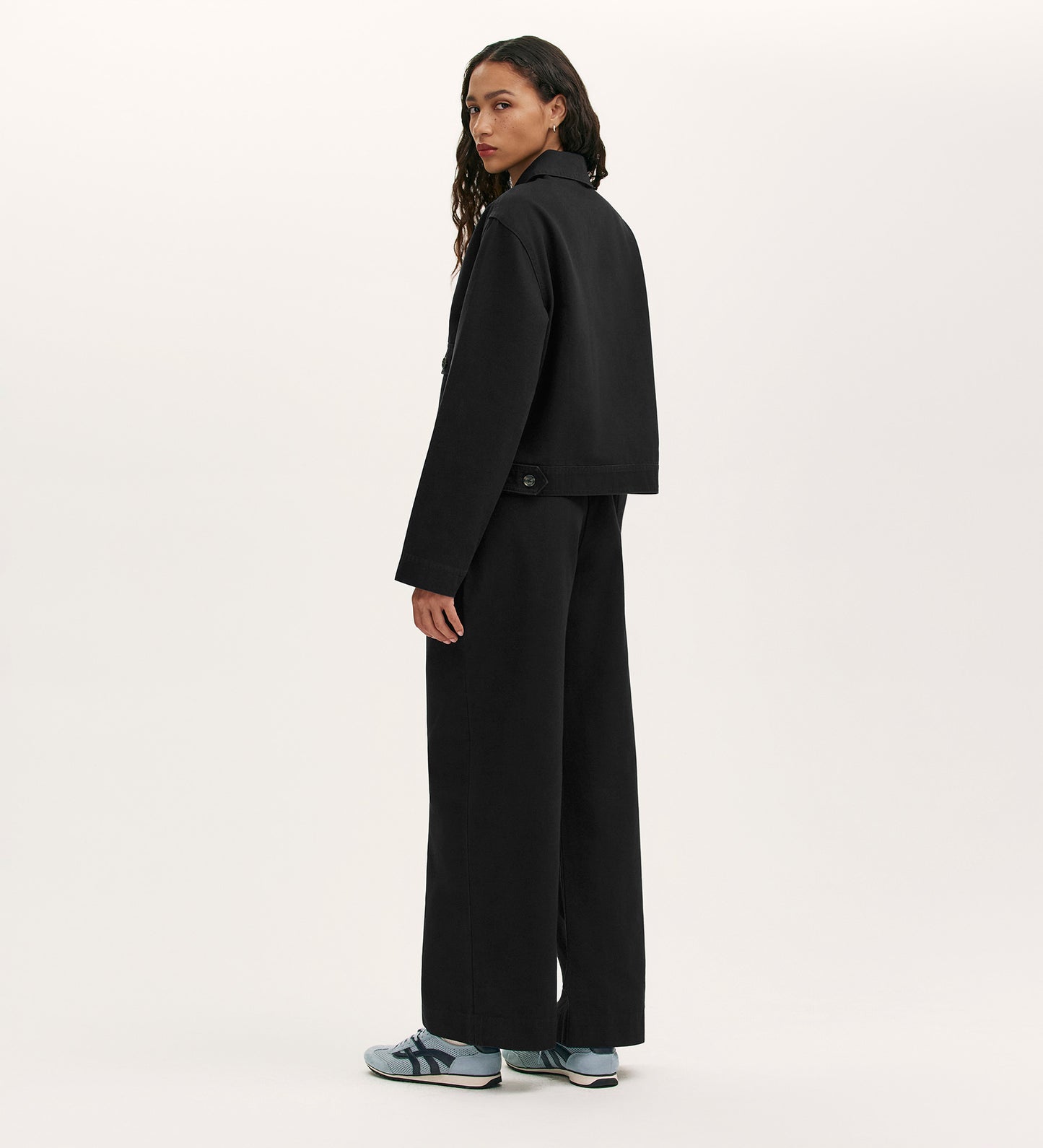 A person with long wavy hair wears the Kelsie Black Canvas Jacket and wide-leg black pants, paired with blue and white sneakers, standing against a plain light background and looking over their shoulder.