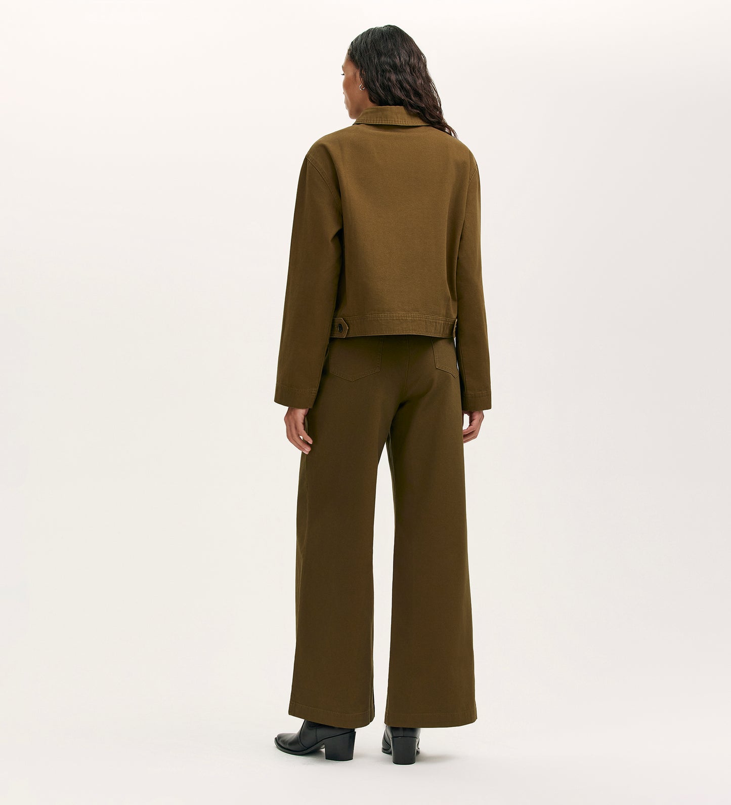 A person with long, wavy hair stands facing away in the Kelsie Khaki Canvas Jacket and matching wide-leg pants, paired with black heeled boots against a plain, light background.