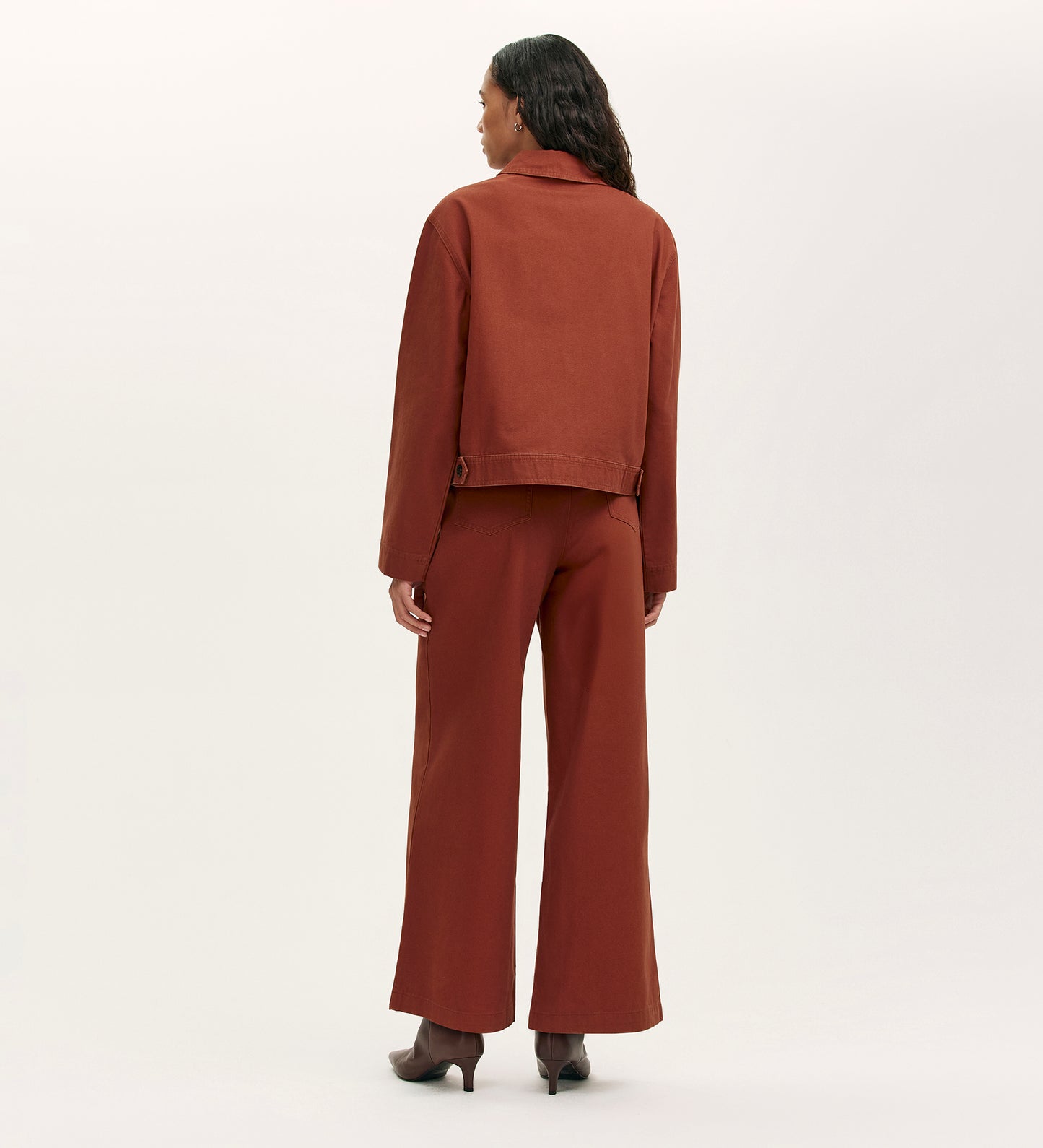 A woman stands with her back to the camera, wearing the Kelsie Terracotta Canvas Jacket and matching wide-leg pants with brown heeled shoes against a plain light background.