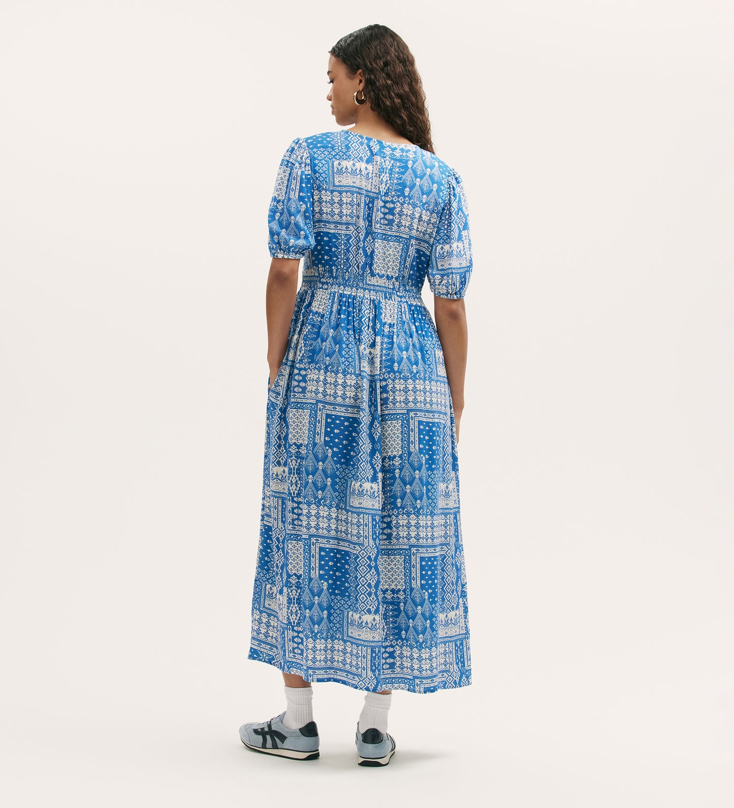 A woman with curly hair faces away, wearing the Kiara Blue Aztec Midi Dress featuring a blue and white pattern, V-neck, side pockets, short sleeves, and paired with white sneakers against a plain light background.