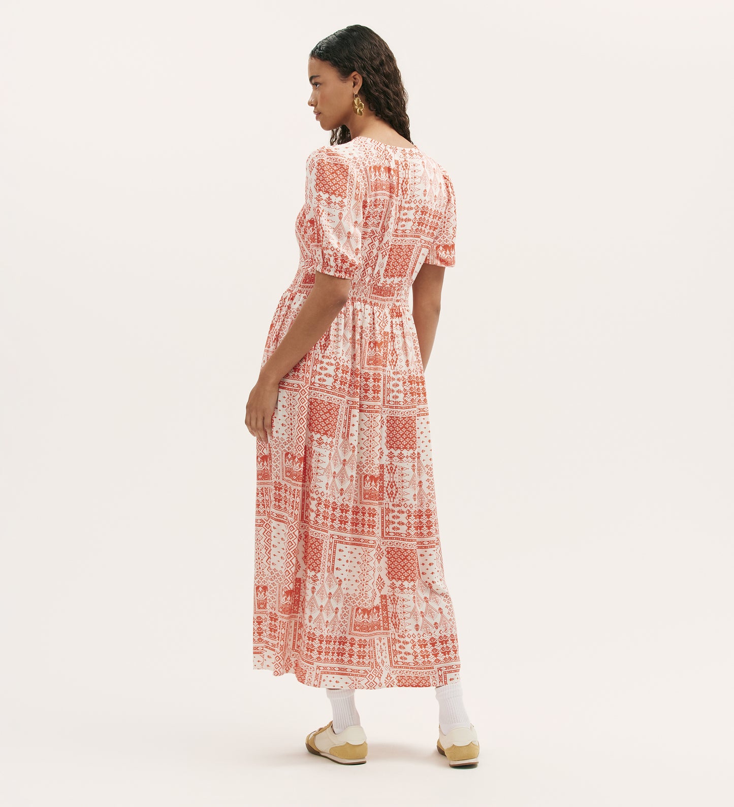 A woman stands facing away in the Kiara Coral Aztec Midi Dress with pockets and short sleeves, paired with white socks and beige shoes. The background is plain and light-colored.