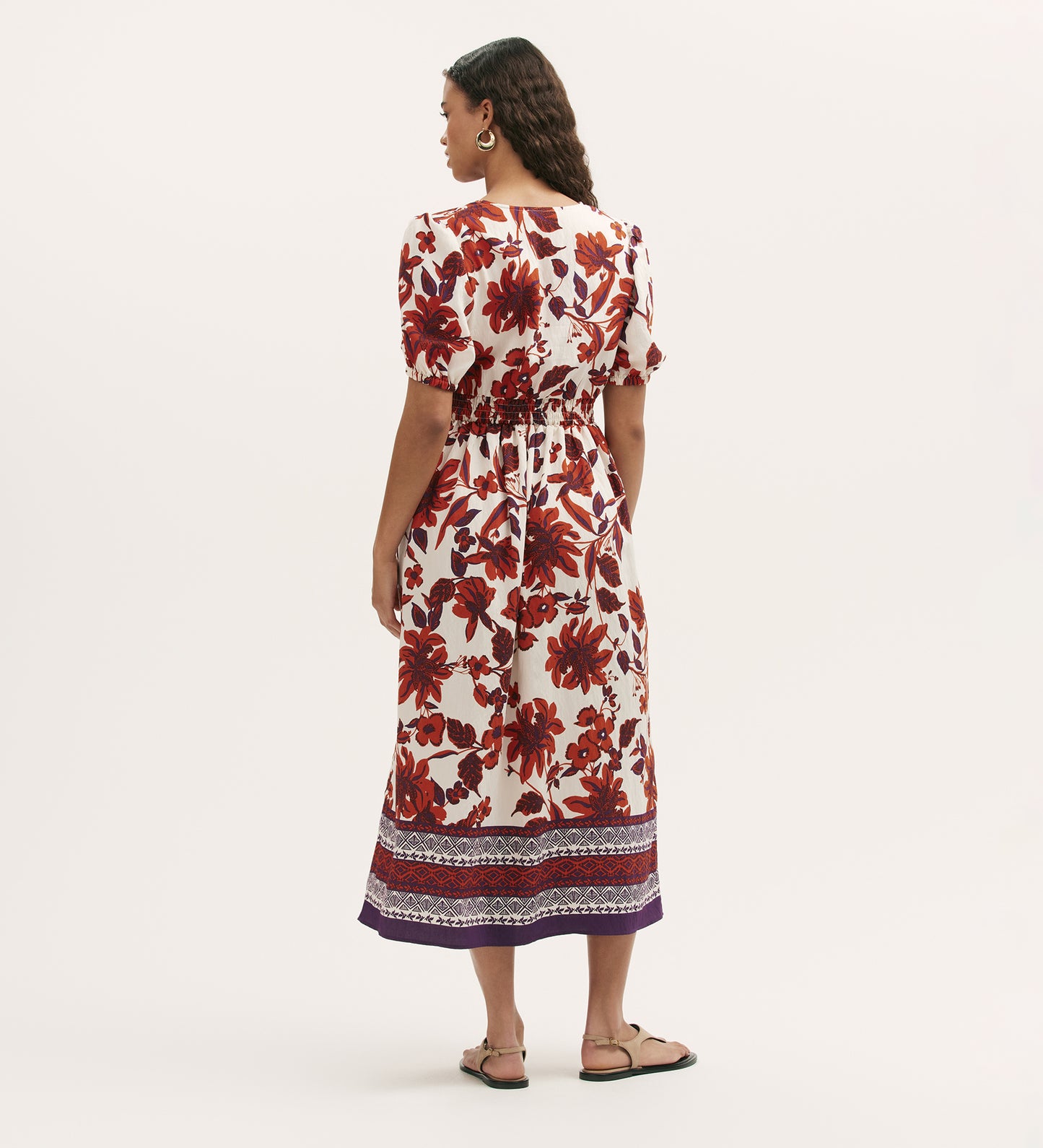 A woman with long wavy hair wears the Kiara Cream Floral Midi Dress, featuring a V-neck, bold red floral prints, shirred waist, side pockets, and a purple patterned hem. She pairs it with sandals and hoop earrings.