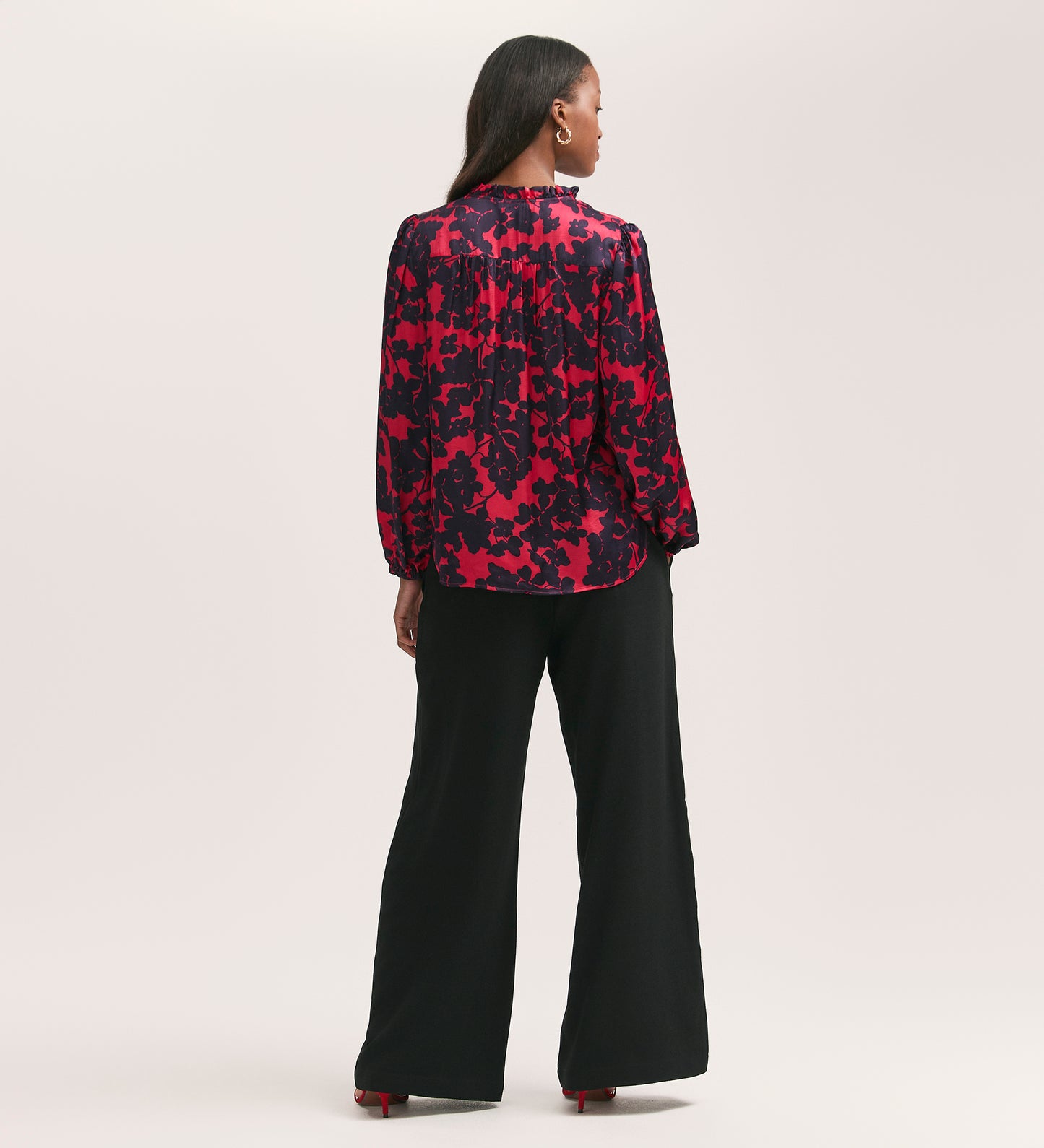 A woman faces away, wearing the relaxed fit Kiya Pink Blooms Crepe Long Sleeve Blouse with a floral pattern and elasticated cuffs, paired with wide-leg black pants and red heeled shoes against a plain light background.