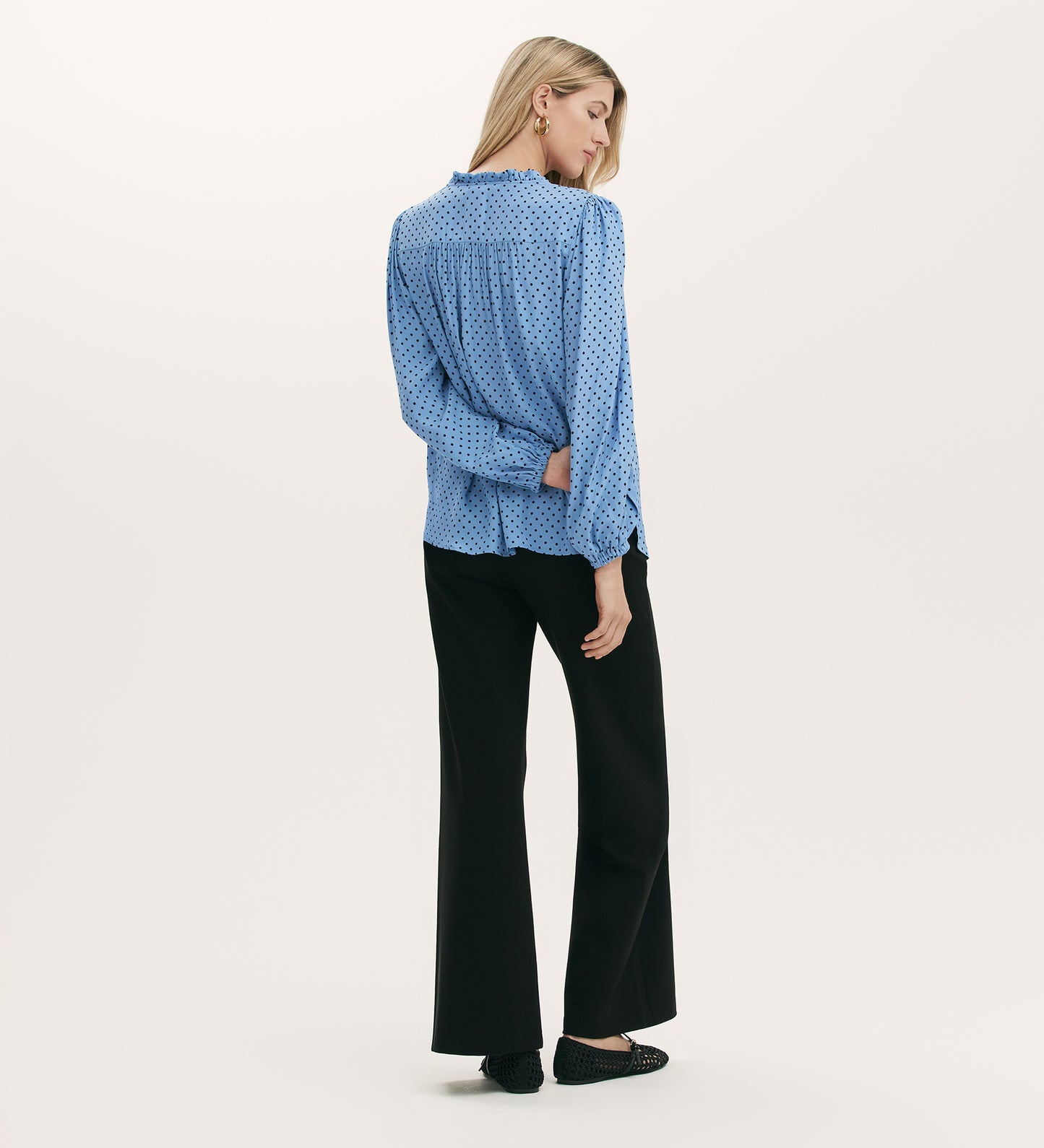 A woman with straight blonde hair poses facing away against a white background, wearing the Kiya Blue Polka Dot Tie Neck Blouse in light blue crepe, paired with black wide-leg pants and black shoes.
