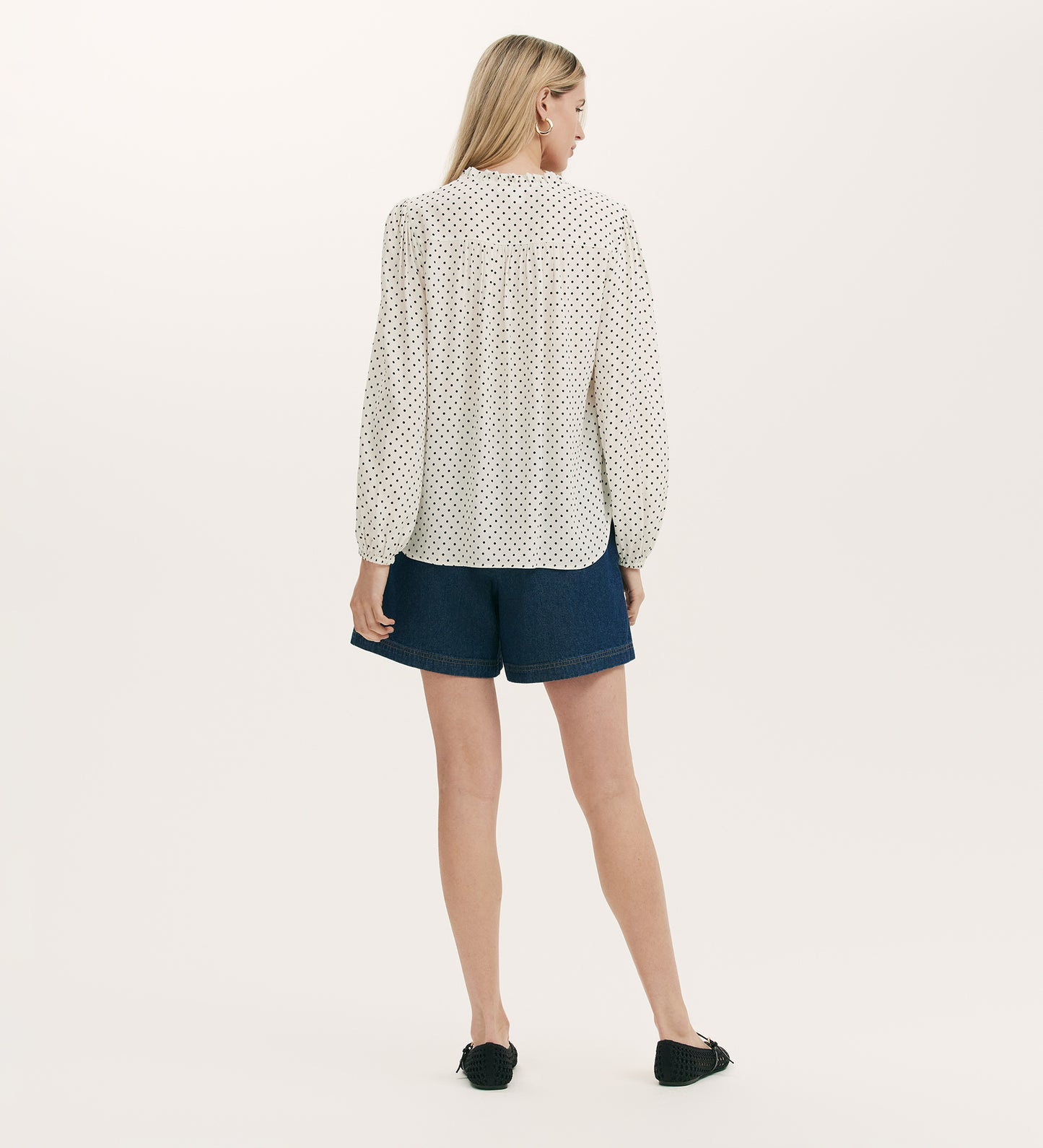A woman faces away, wearing the Kiya Ivory Polka Dot Tie Neck Blouse, paired with dark blue denim shorts and black slip-on shoes against a plain light background.