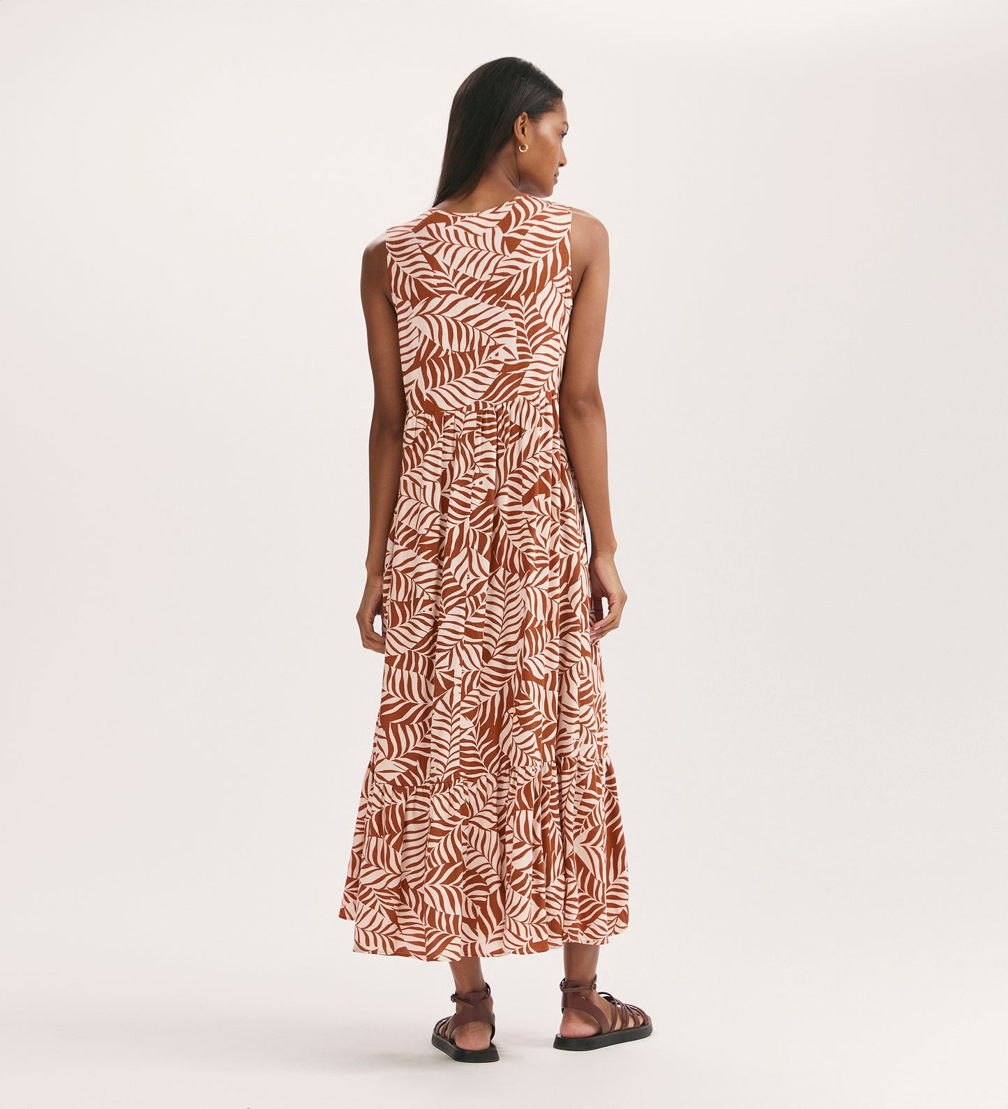 A woman faces away in the Larissa Tan Leaf Tie Front Midi Dress, featuring a sleeveless cut, tan leaf print, functional pockets, and paired with black sandals against a plain light background.