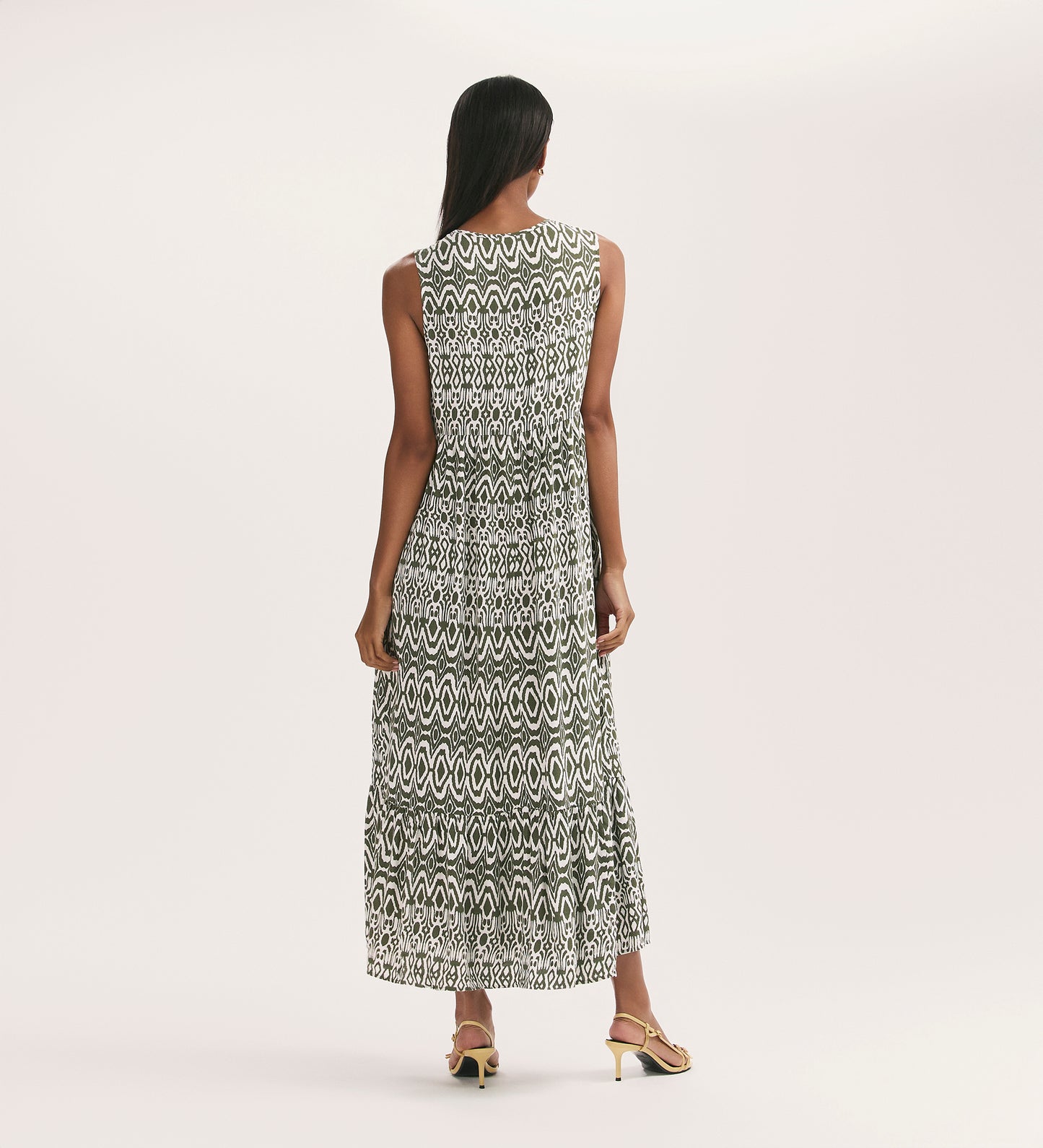 A woman with long dark hair faces away, wearing the Larissa Khaki Tribal Tie Front Midi Dress featuring a black and white geometric pattern, functional pockets, and open-toe heels against a plain light background.