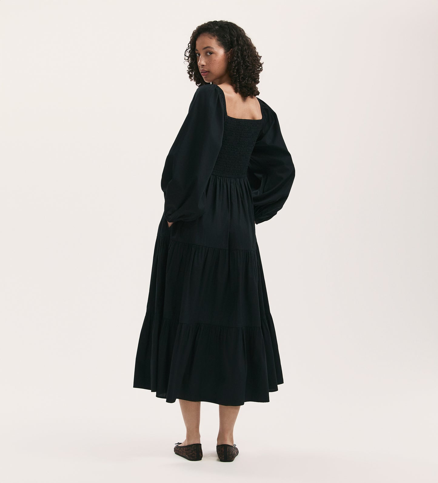 A woman with curly hair wears the Lauretta Black Cotton Midi Tiered Dress with long sleeves, a square neckline, tiered skirt, and smocked back. She stands facing away, glancing over her shoulder in black flats against a plain background.
