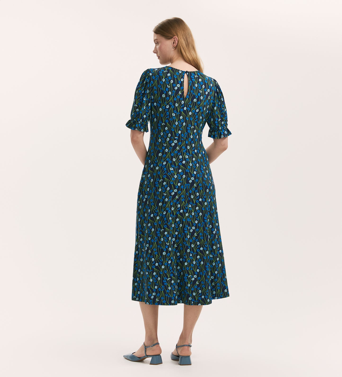 A woman with light brown hair, seen from behind, wears the Lenisha Blue Flower Jersey Midi Tea Dress with short puff sleeves and blue low-heeled shoes, set against a plain light background.
