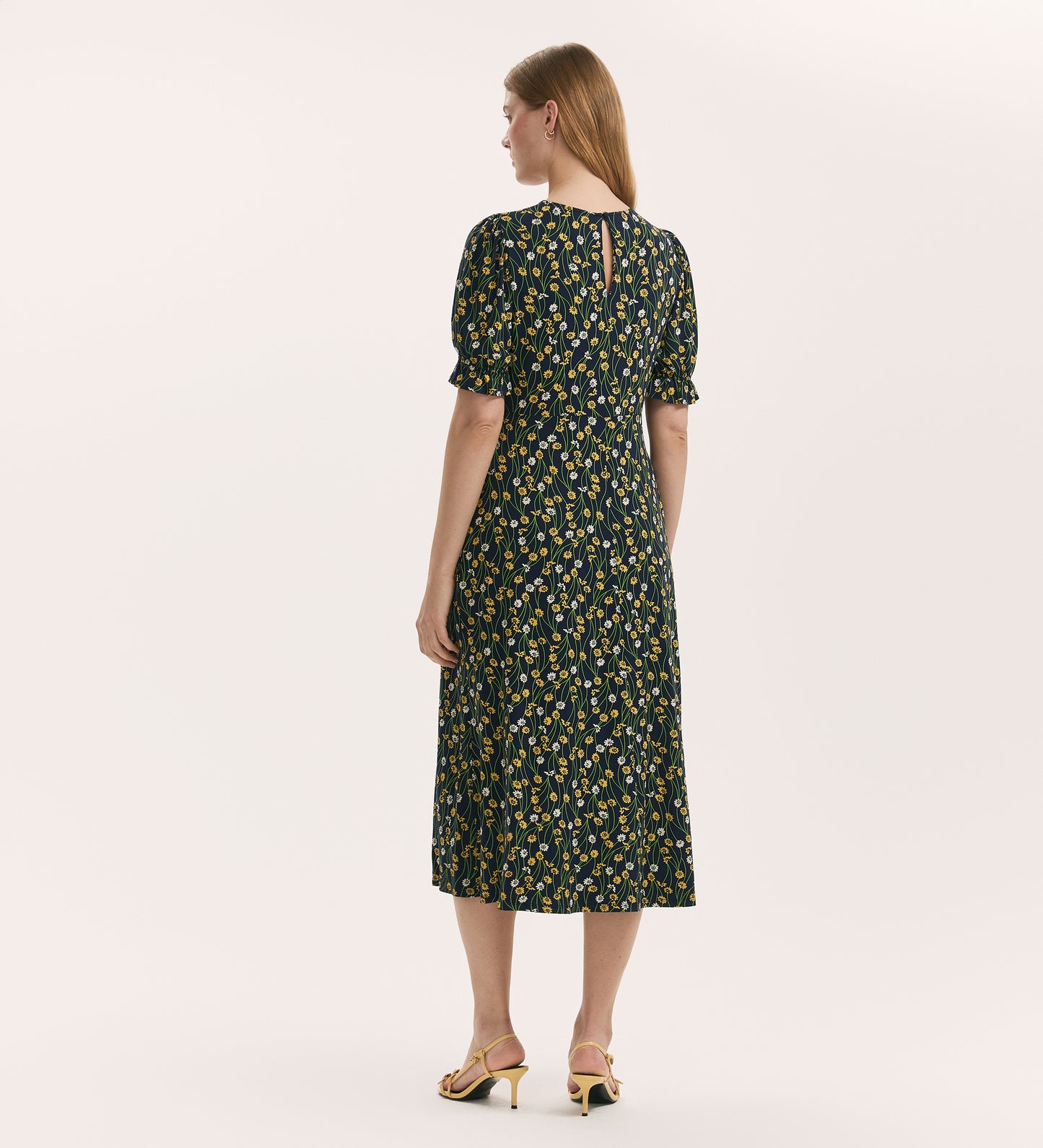 A woman stands with her back to the camera, wearing the Lenisha Yellow Flower Jersey Midi Tea Dress and yellow heeled sandals against a plain light background.