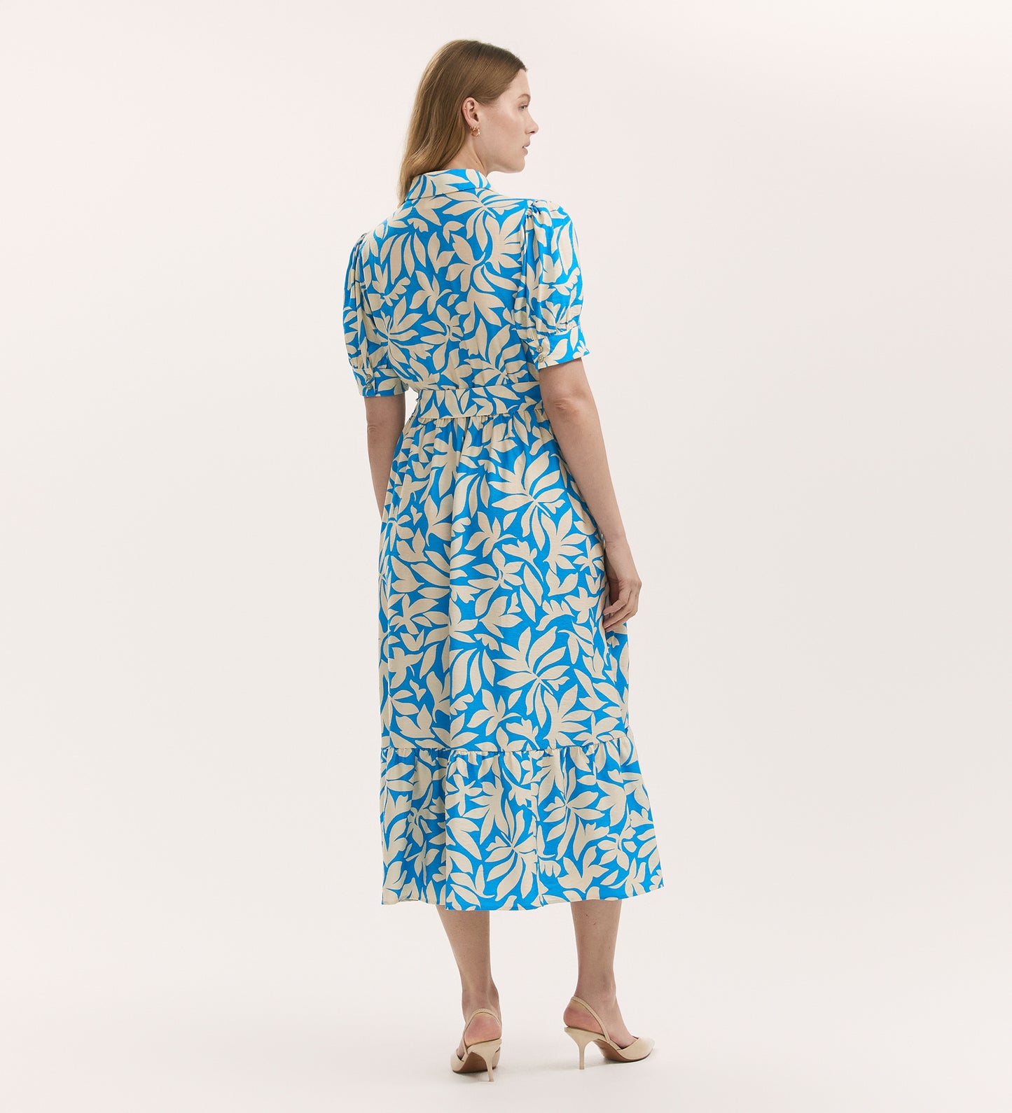 A woman stands facing away in the Liana Blue Leaf Midi Tiered Dress—featuring a blue and yellow leaf print, tiered midi-length skirt, short sleeves, and removable tie belt—paired with beige heeled sandals against a white background.