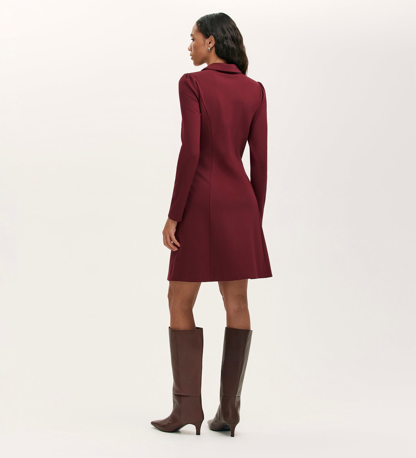 A woman stands with her back to the camera, dressed in the Lea Burgundy Ponte Jersey Short Dress and tall brown heeled boots against a plain light background.