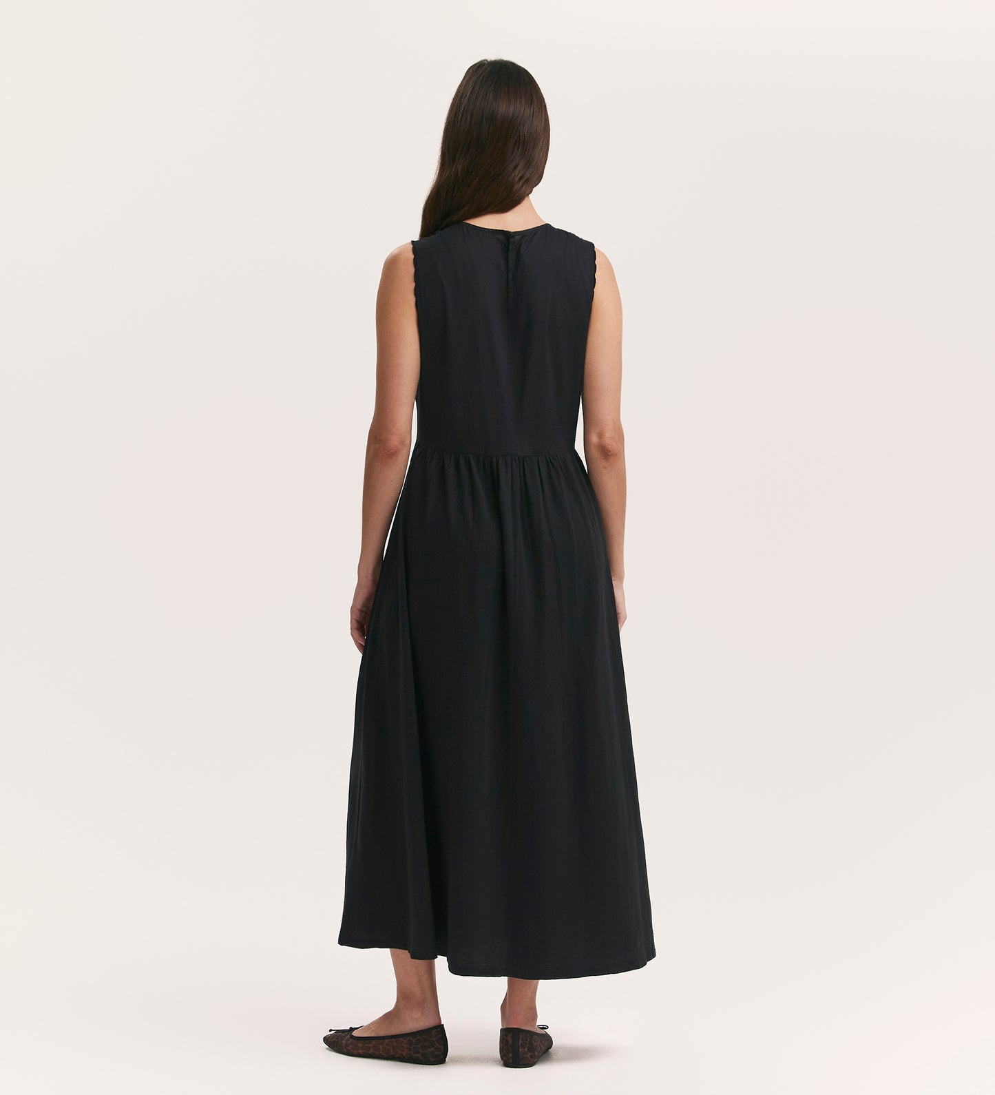 A woman with long brown hair, seen from behind, wears the Liberte Black Cotton Sleeveless Midi Dress and black flats, standing against a plain light background.