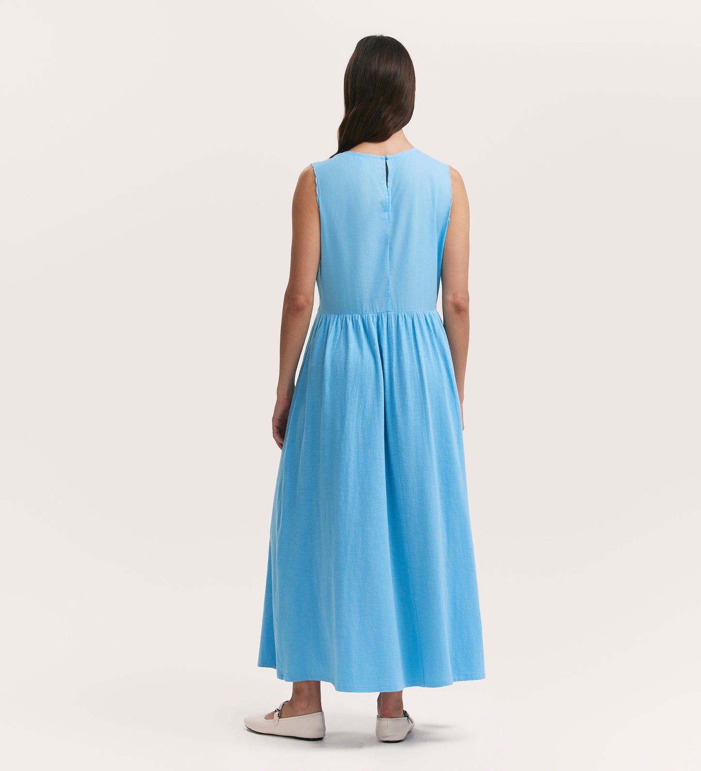 A woman with long brown hair, wearing the Liberte Light Blue Cotton Sleeveless Midi Dress and white flats, stands facing away against a plain light background.