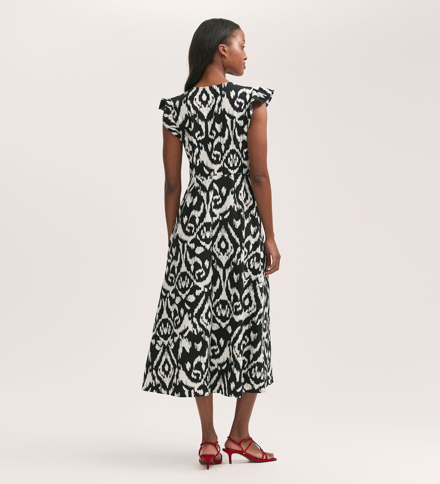 A woman faces away in the Liza Black Ikat Crepe Midi Dress, featuring short flutter sleeves and crease-free fabric, paired with red strappy sandals against a plain light background.