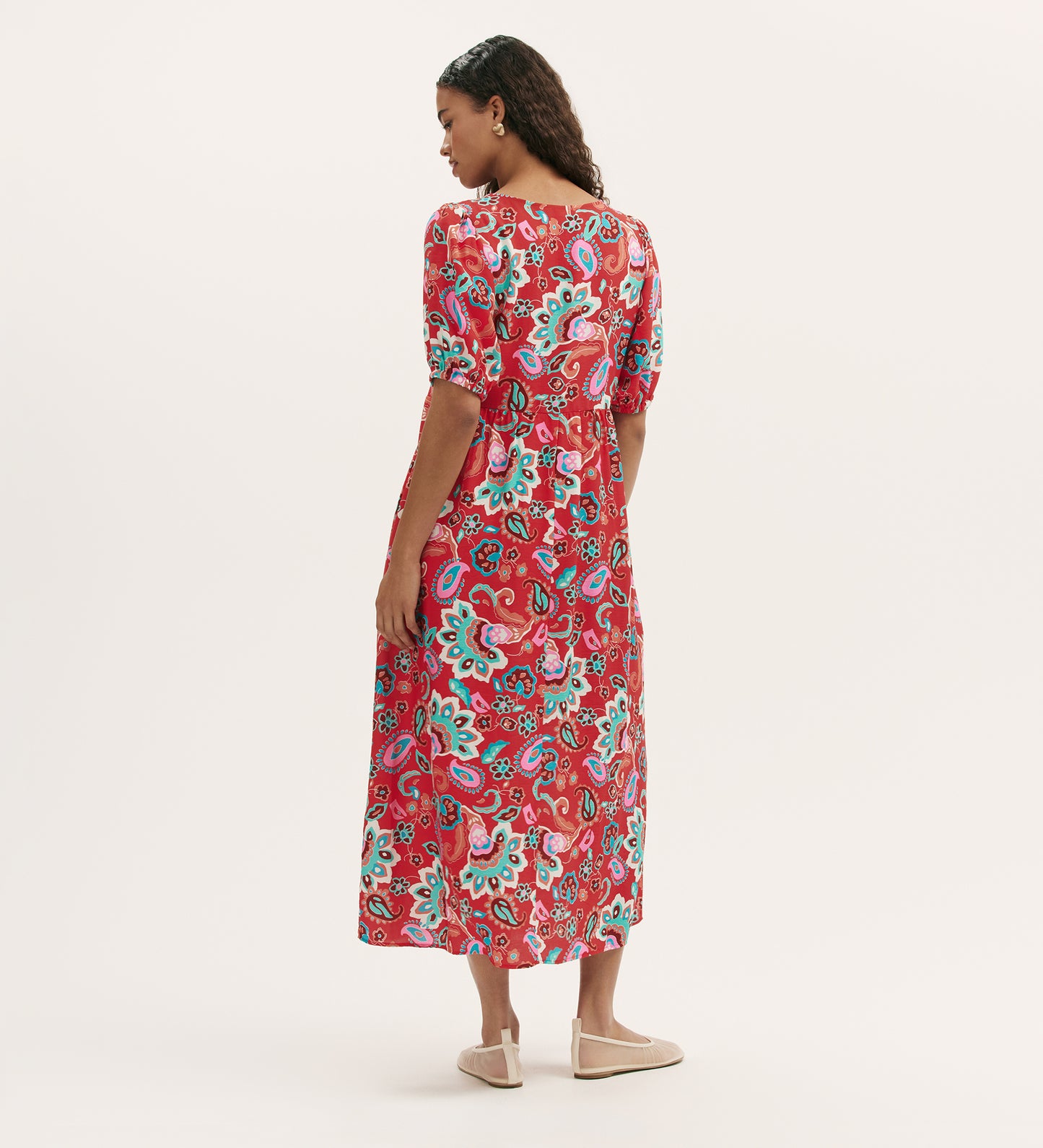 A woman faces away, wearing the Logan Red Paisley Midi Dress featuring a vibrant paisley print and elbow-length sleeves. The relaxed-fit, ankle-length dress is paired with beige flats against a plain white background.