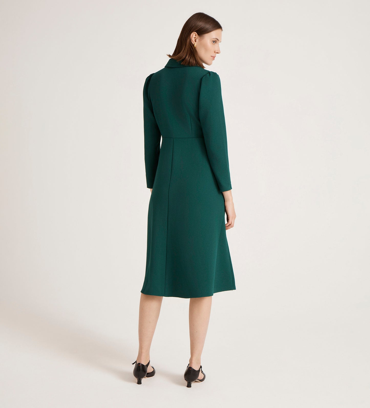 A woman faces away in the Lucia Bottle Green Crepe Midi Dress, featuring a classic collar, paired with black heeled sandals against a plain light background.