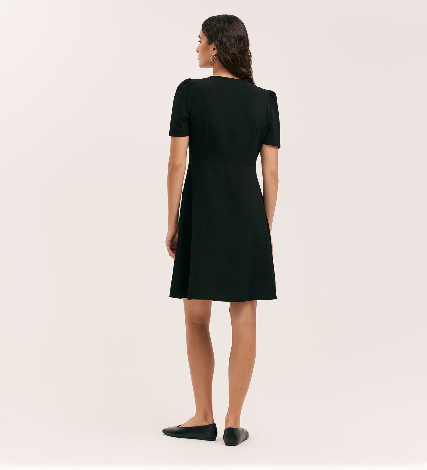 A woman with loose, wavy brown hair faces away, wearing the Lucille Black Crepe Short Dress with a tailored silhouette and black flats, against a plain light background.