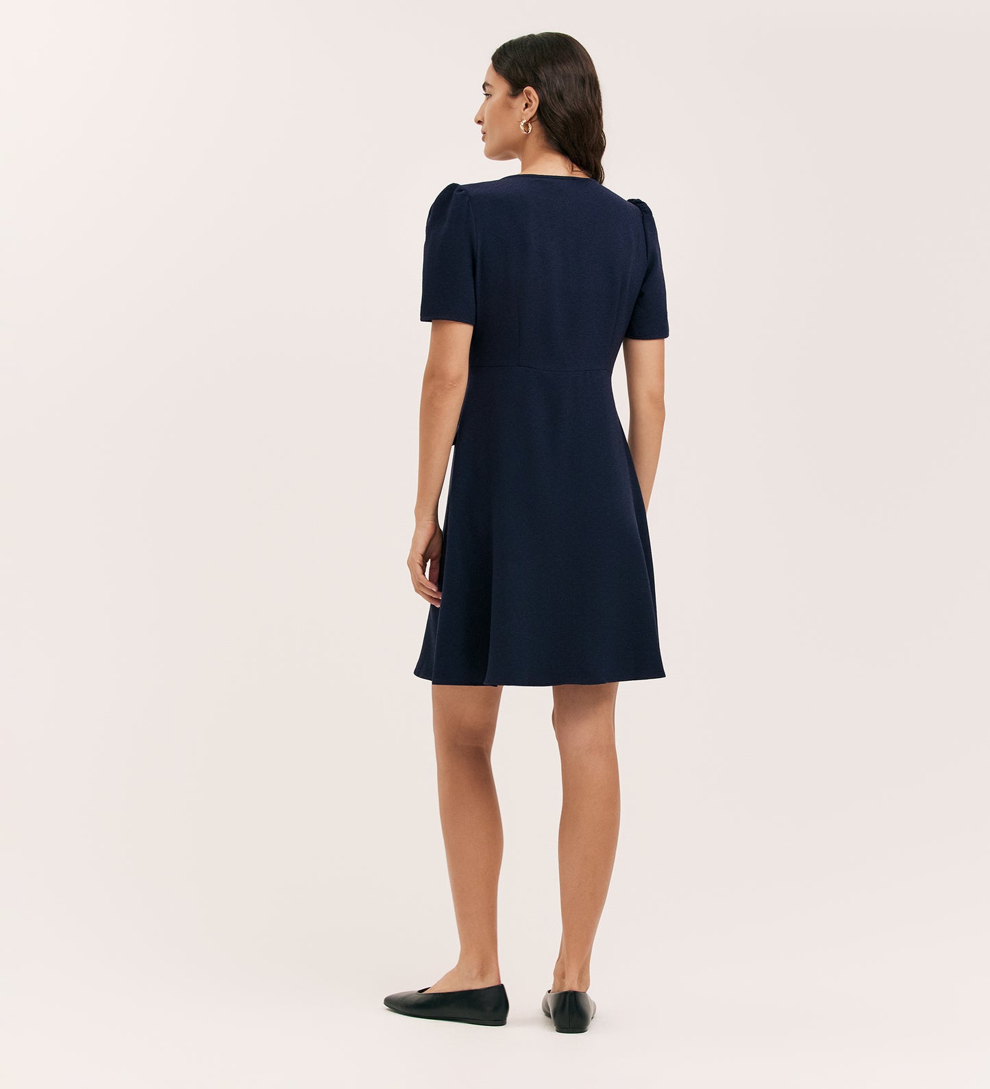 A woman stands with her back to the camera in the Lucille Navy Crepe Short Dress, featuring a tailored silhouette and knee-length cut, paired with black flat shoes against a plain light background.