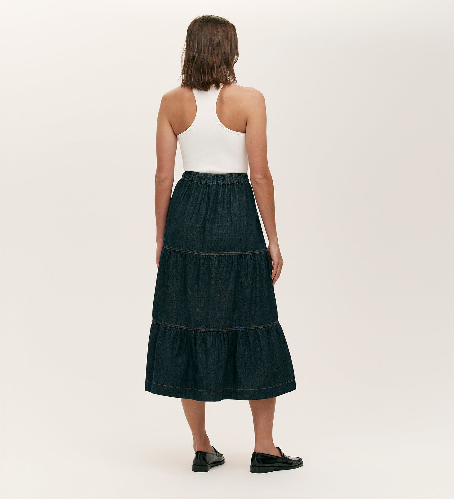 Someone stands facing away, wearing a white racerback tank top, black slip-on shoes, and the Lulu Indigo Denim Midi Skirt featuring a dark wash, tiered silhouette, and elasticated waistband, set against a plain light background.
