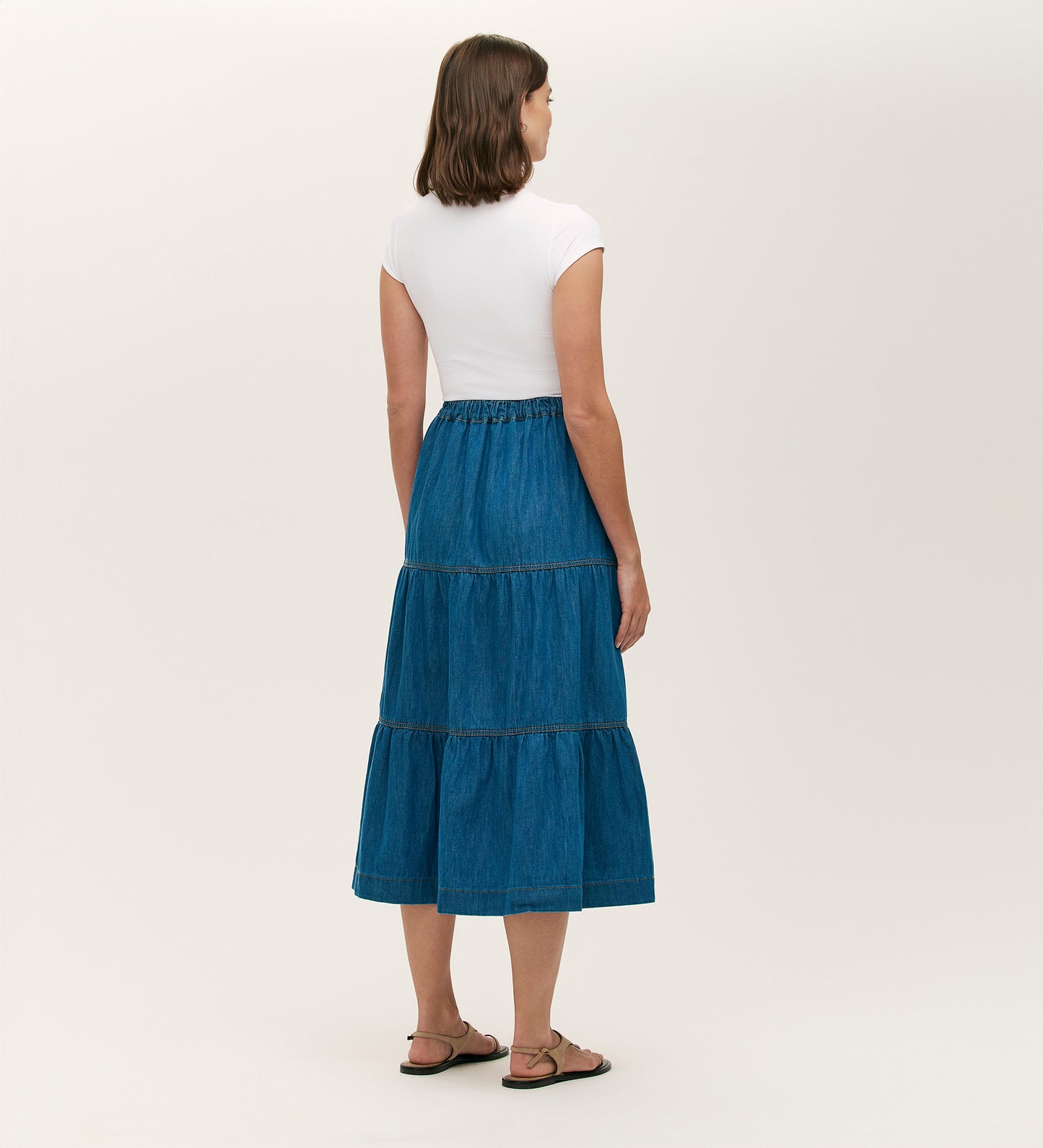 A woman faces away, wearing a white short-sleeve top and the Lulu Light Blue Denim Midi Skirt with an elasticated waistband and tiered silhouette, paired with black sandals against a plain light background.