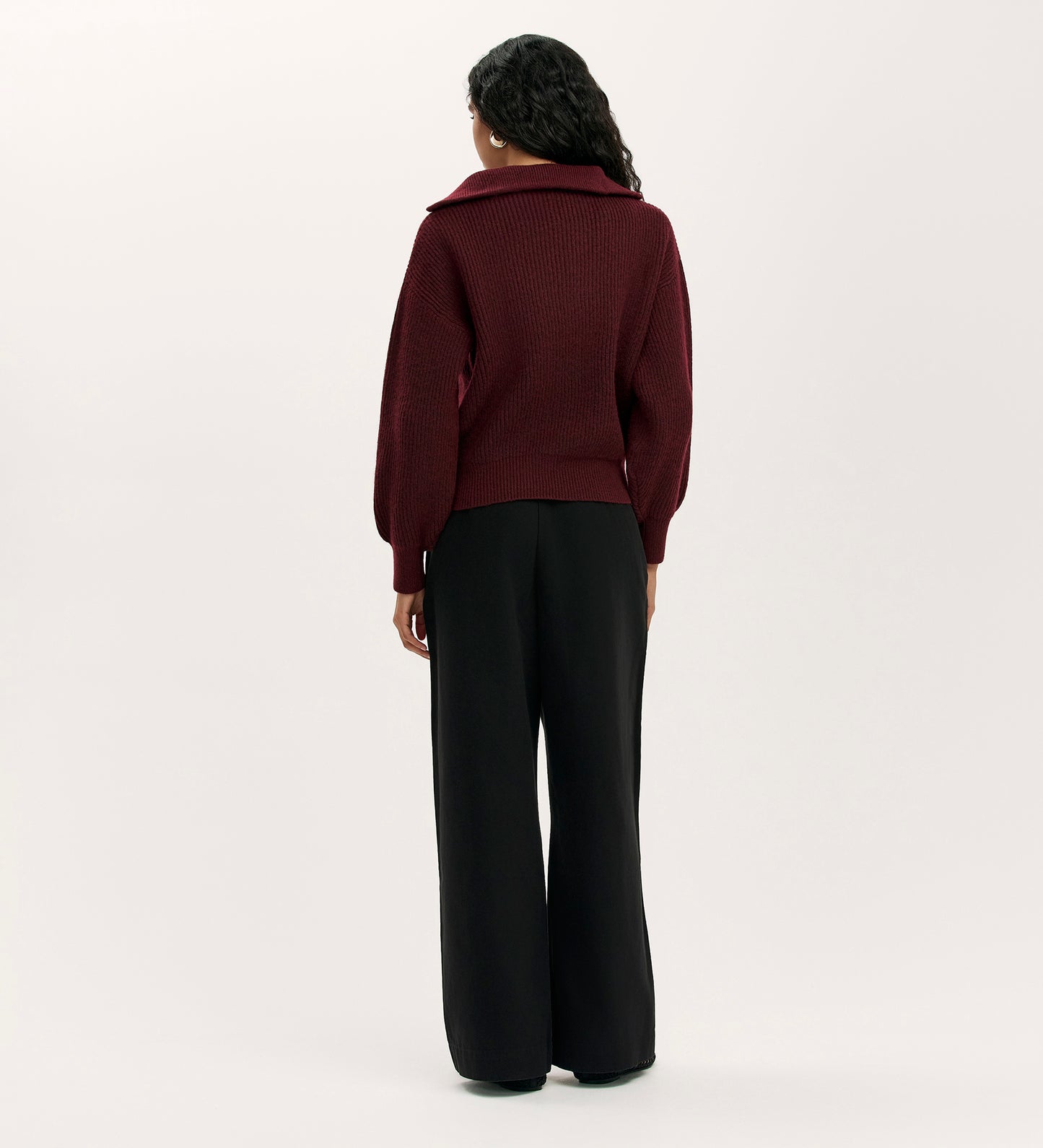A person with long, wavy hair faces away, wearing the Maddy Burgundy Knitted Jumper with a wide collar and black wide-leg pants against a plain light background.