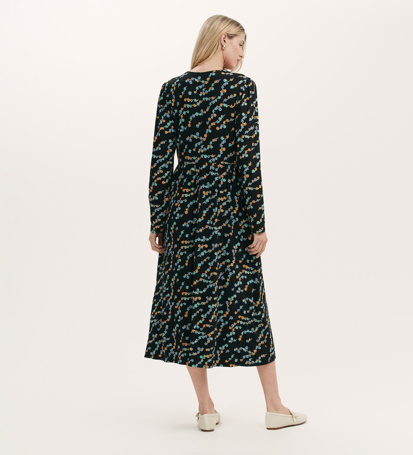 A woman with long blonde hair, seen from behind, wears the Madelyn Black Rose Bud Midi Dress with a colorful abstract pattern and light shoes, standing against a plain light background.