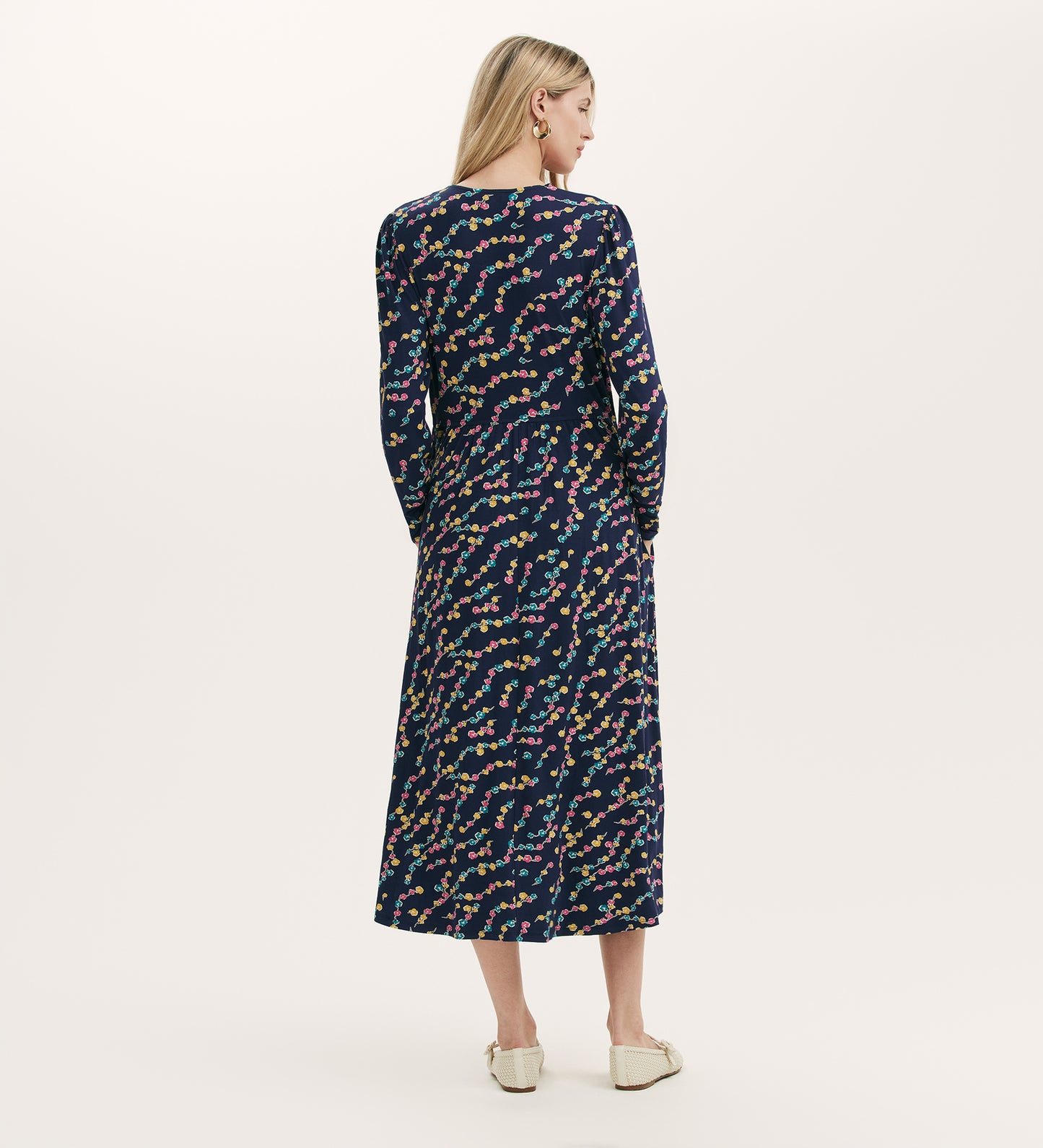 A woman with long blonde hair, facing away against a light background, wears the Madelyn Navy Rose Bud Midi Dress—featuring a colorful abstract pattern and V-neckline—with cream shoes.