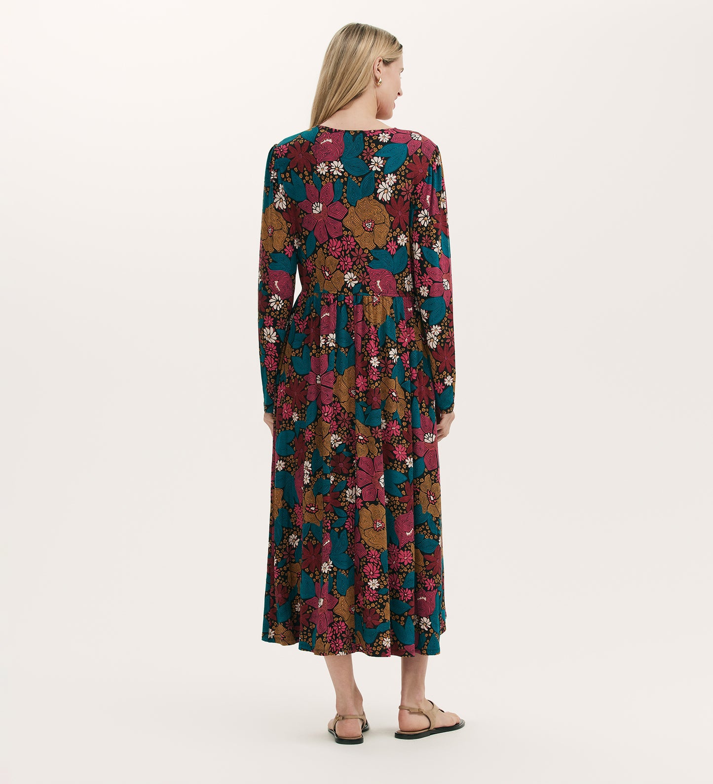 A woman with long blonde hair, seen from behind, wears the Madelyn Pink Floral Midi Dress featuring long sleeves and a V-neckline with sandals, standing against a plain light background.