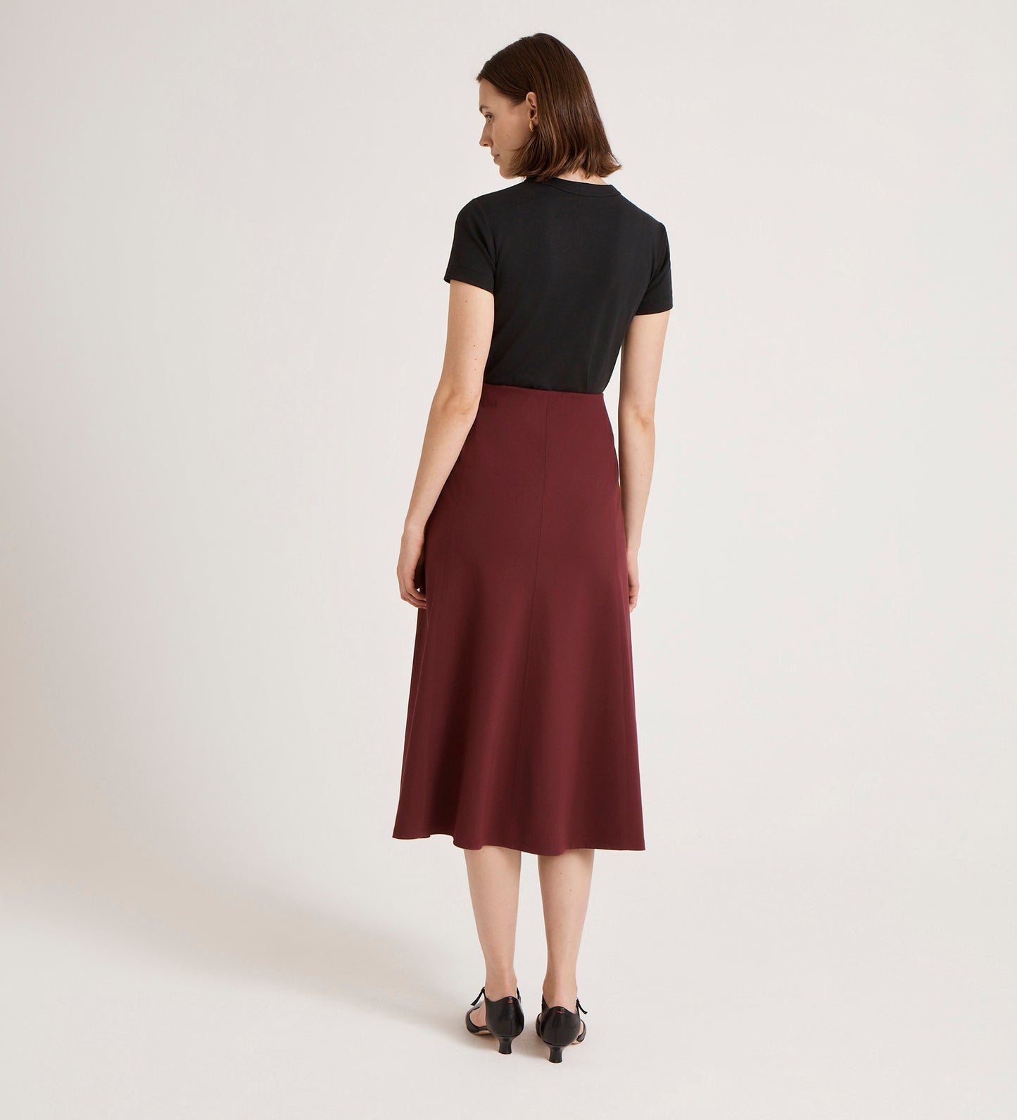 A woman stands facing away from the camera in a black short-sleeve top and the Mae Burgundy Ponte Jersey Midi Skirt with an elasticated waistband, paired with black heeled sandals, against a plain white background.