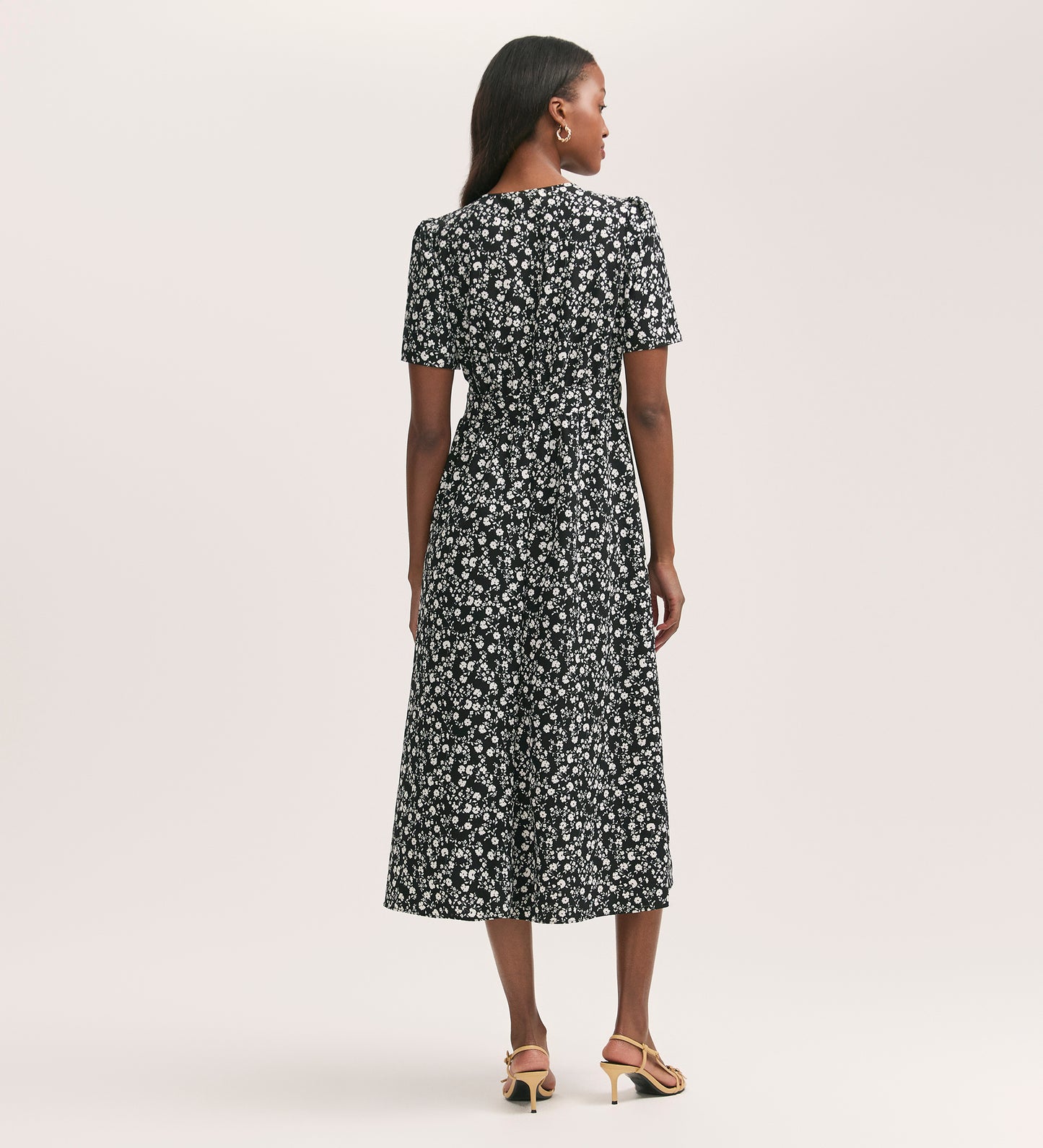 A woman with medium-length hair and tan heeled sandals stands against a plain light background, wearing the Mairead Black Ditsy Crepe Midi Tea Dress in a flattering fit-and-flare style with short sleeves.