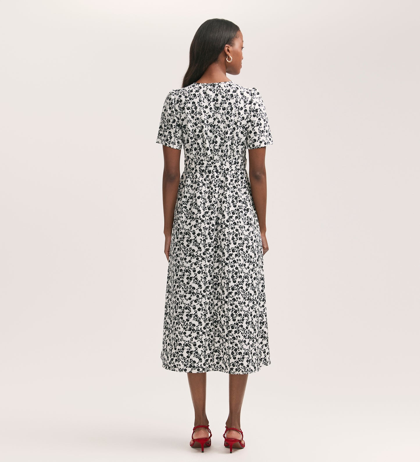 A woman faces away, wearing the Mairead Ivory Ditsy Crepe Midi Tea Dress in a fit and flare style with short sleeves and red heeled sandals, set against a plain light-colored background.