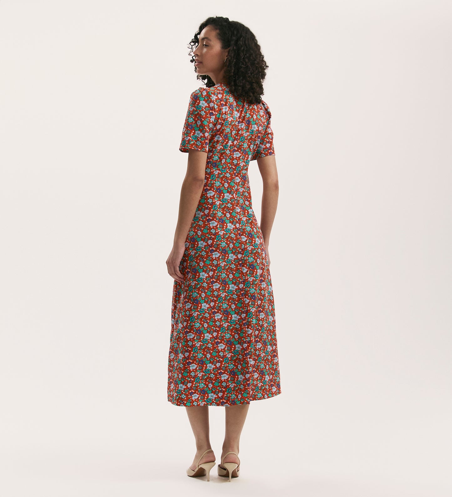 A woman with curly hair, facing away, wears the Marie Red Ditsy Crepe Midi Tea Dress with beige heels against a plain white background.