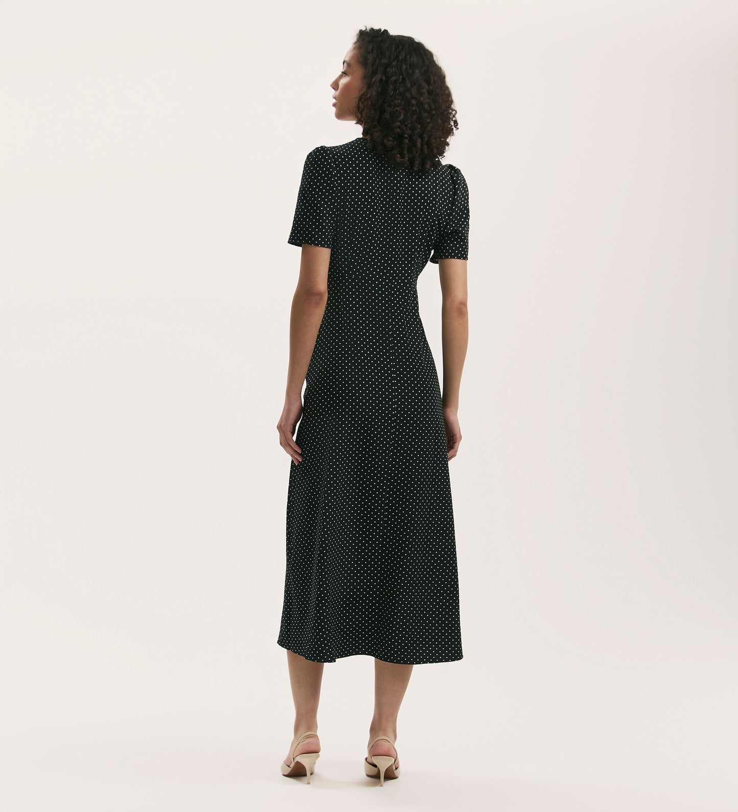 A woman with curly hair, seen from behind, wears the Marie Black Polka Dot Crepe Midi Tea Dress and light open-toe heels against a plain light background.