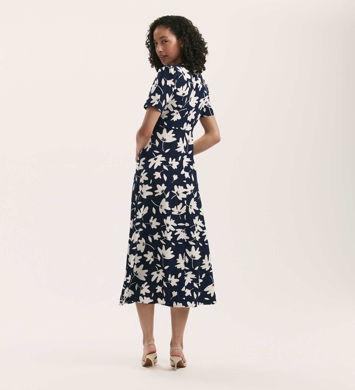 A woman with curly hair models the Marie Navy Leaves Crepe Midi Tea Dress, featuring a white floral pattern and button details. She wears light heeled sandals and looks over her shoulder against a plain background.