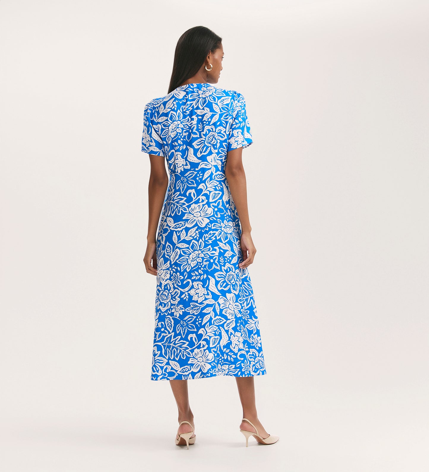 A woman stands facing away, wearing the Martina Blue Flower Crepe Midi Dress—a bright blue, short-sleeved midi dress with a bold white floral print. She pairs it with beige low-heeled sandals, and the crease-free fabric looks fresh against a plain background.