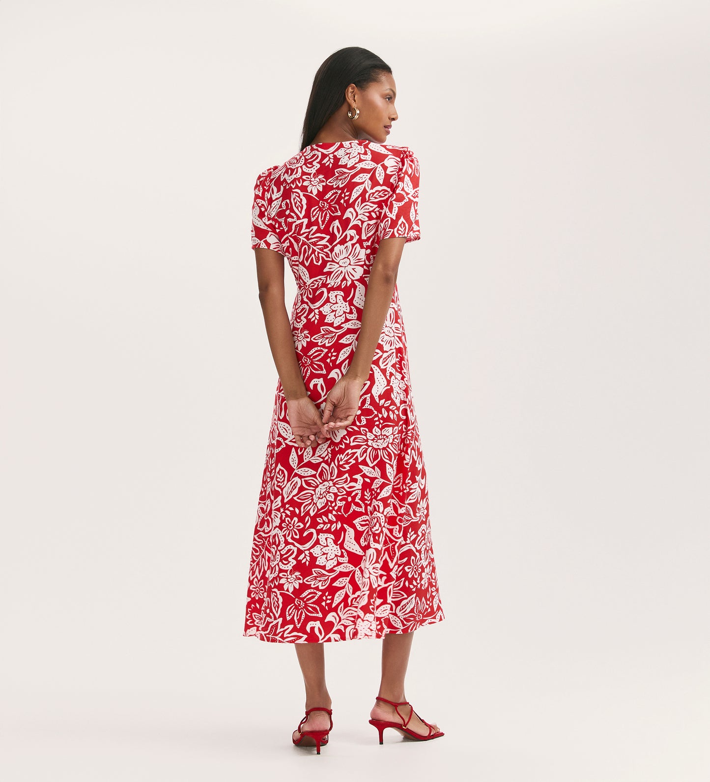 A woman stands with her back to the camera, wearing the Martina Red Flower Crepe Midi Dress with a button-down front and red heeled sandals, hands clasped behind her against a plain light background.