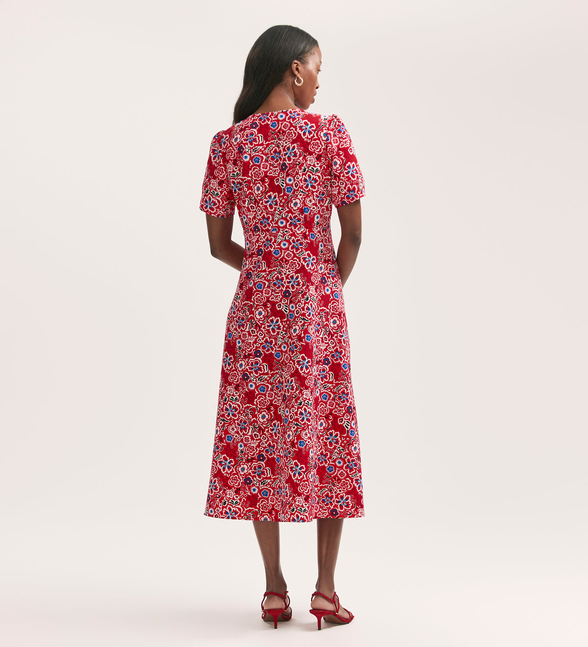 A woman faces away, wearing the Martina Red Flower Crepe Midi Dress with a button-down front and short sleeves, styled with red heels. The crease-free crepe fabric drapes elegantly against a plain, light-colored background.